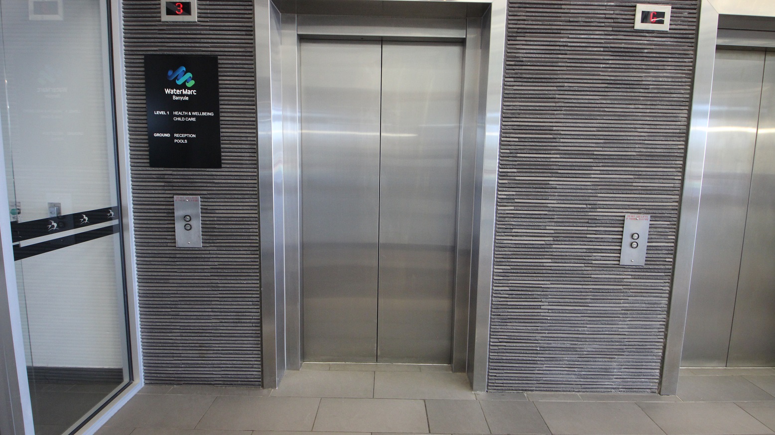Two lift doors are visible, with the one on the left partially open, revealing a mirrored interior. The central lift door is closed, with a metallic finish. Both lifts are set within a wall covered in horizontal grey tiles. Above the left lift is a sign for "WaterMarc Banyule" with floor information. Each lift has a control panel with buttons. The floor is tiled in light grey, and there is a small display above each lift showing the floor number. Two lift doors are visible, with the one on the left partially open, revealing a mirrored interior. The central lift door is closed, with a metallic finish. Both lifts are set within a wall covered in horizontal grey tiles. Above the left lift is a sign for "WaterMarc Banyule" with floor information. Each lift has a control panel with buttons. The floor is tiled in light grey, and there is a small display above each lift showing the floor number.