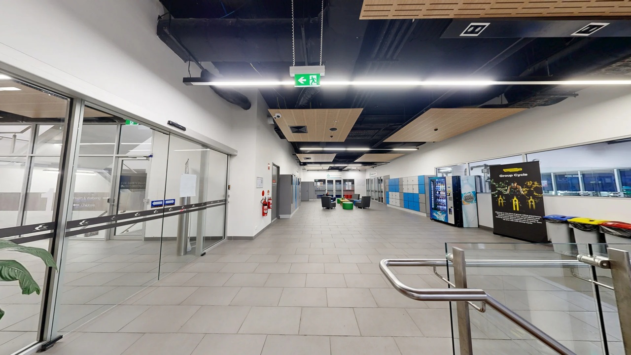 A spacious indoor area with a grey tiled floor and a ceiling featuring black and wooden panels. On the left, there are large glass doors with a plant visible through them. In the centre, there are seating arrangements with chairs and tables. On the right, vending machines and a display board are present, along with a row of colourful bins. An exit sign hangs from the ceiling, and a metal barrier is in the foreground. A spacious indoor area with a grey tiled floor and a ceiling featuring black and wooden panels. On the left, there are large glass doors with a plant visible through them. In the centre, there are seating arrangements with chairs and tables. On the right, vending machines and a display board are present, along with a row of colourful bins. An exit sign hangs from the ceiling, and a metal barrier is in the foreground.