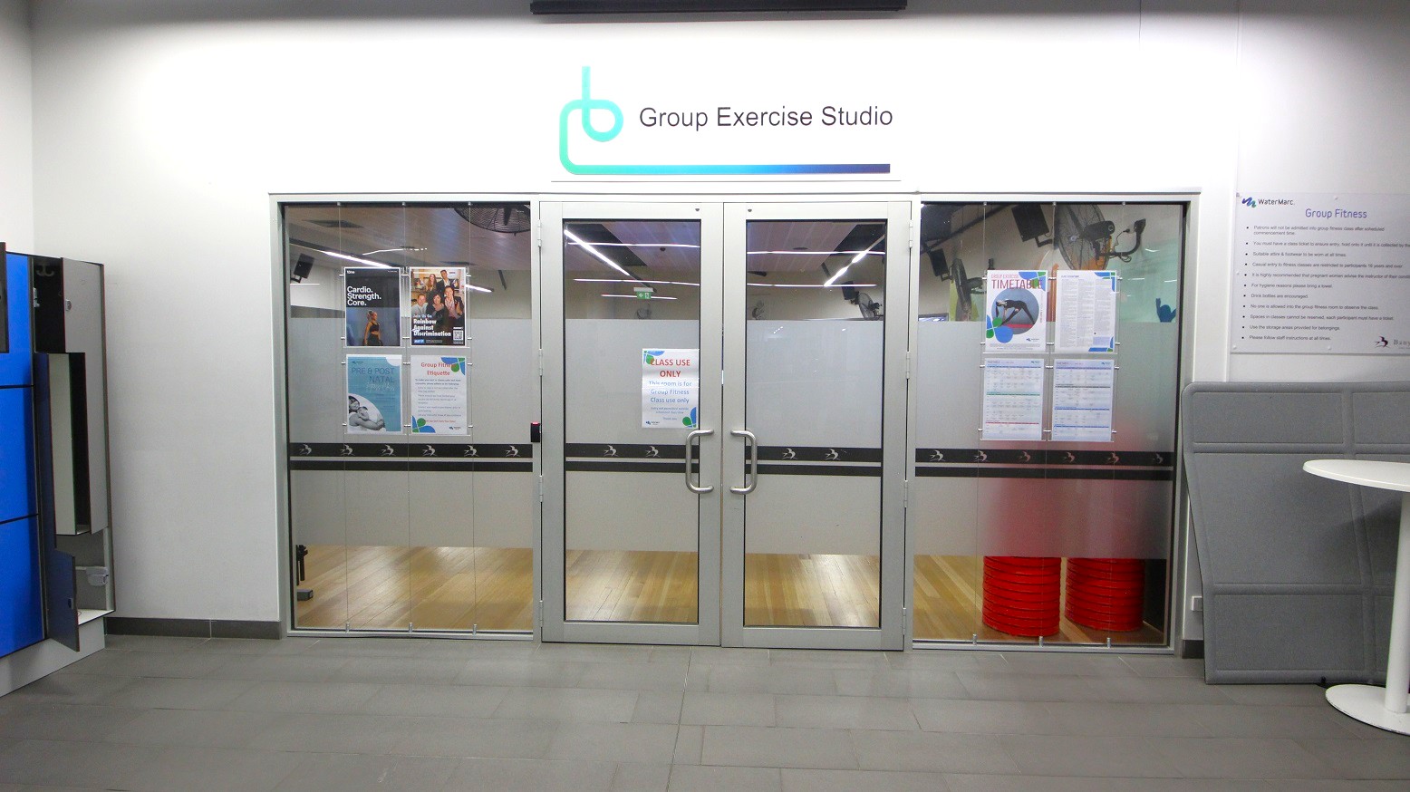 Glass doors leading to a group exercise studio, with posters and notices displayed on them. Above the doors, a sign reads "Group Exercise Studio" with a logo. The floor inside is wooden, while the exterior floor is tiled. On the left, there are blue lockers, and on the right, a white round table and a grey partition. The ceiling has fluorescent lights. Glass doors leading to a group exercise studio, with posters and notices displayed on them. Above the doors, a sign reads "Group Exercise Studio" with a logo. The floor inside is wooden, while the exterior floor is tiled. On the left, there are blue lockers, and on the right, a white round table and a grey partition. The ceiling has fluorescent lights.