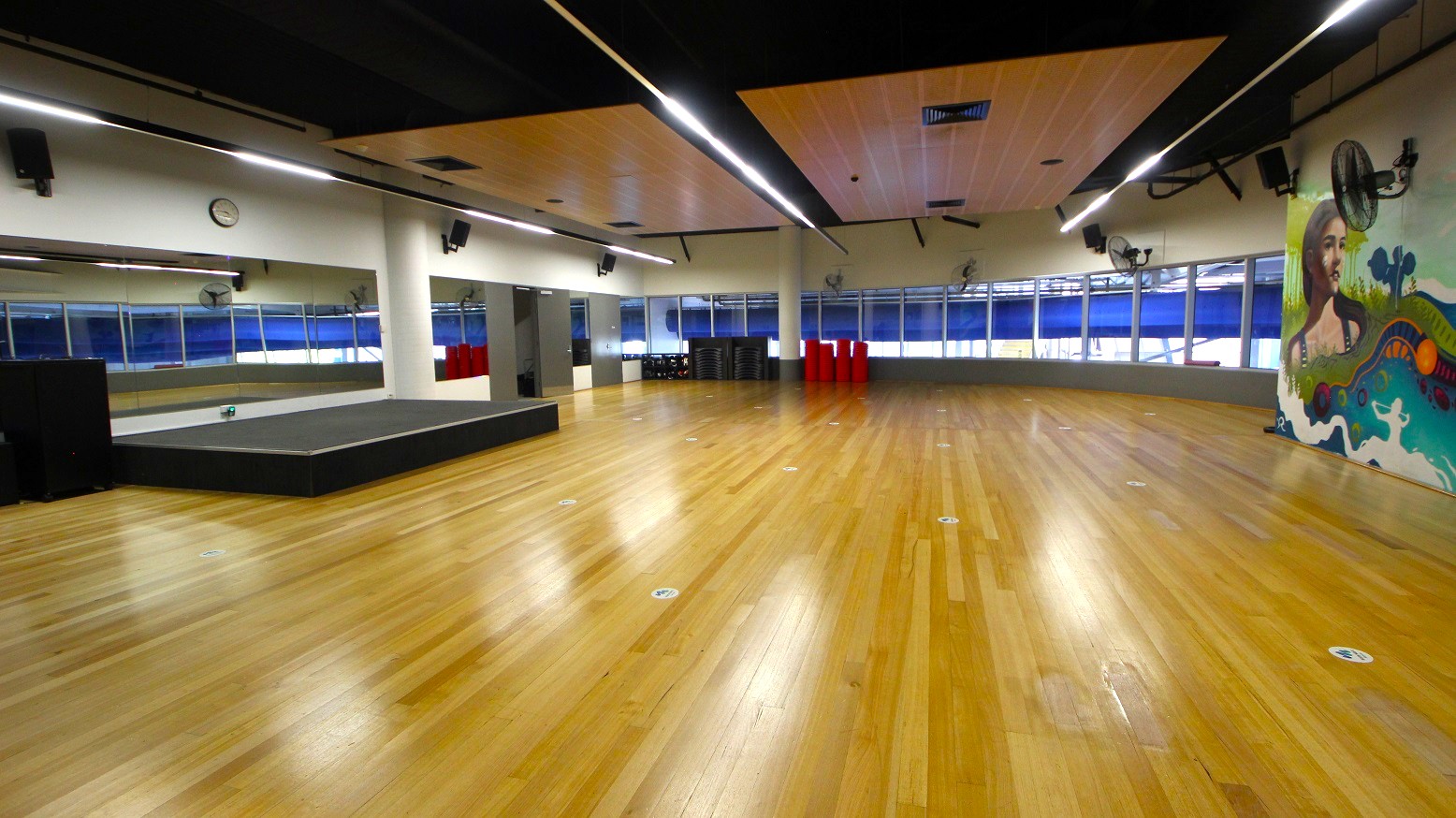 A spacious room with a polished wooden floor and a high ceiling featuring recessed lighting. On the left, there's a large mirror and a small stage. The back wall has large windows with blue blinds. On the right, a colourful mural is visible. Several red cylindrical objects and stacked black equipment are against the back wall. Ceiling fans are mounted on the walls. A spacious room with a polished wooden floor and a high ceiling featuring recessed lighting. On the left, there's a large mirror and a small stage. The back wall has large windows with blue blinds. On the right, a colourful mural is visible. Several red cylindrical objects and stacked black equipment are against the back wall. Ceiling fans are mounted on the walls.