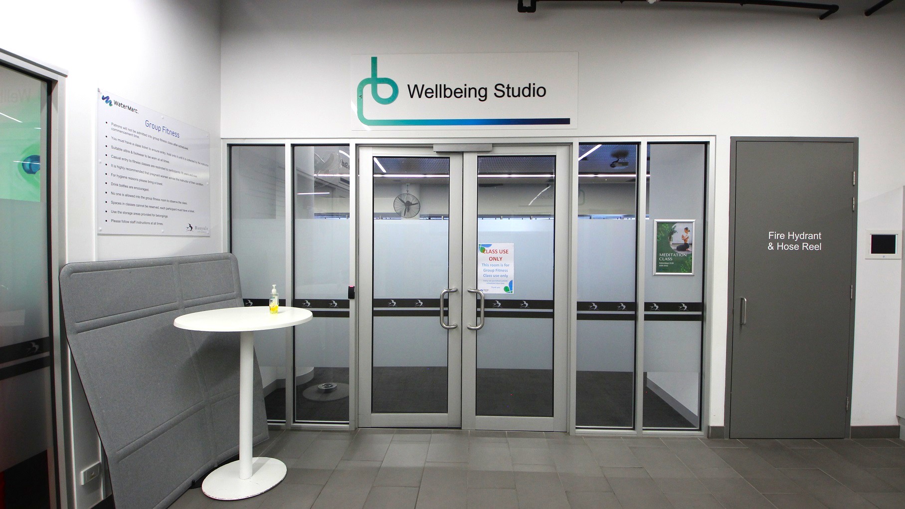 Entrance to a "Wellbeing Studio" with glass double doors in the centre, featuring a sign above. To the right, a grey door marked "Fire Hydrant & Hose Reel". On the left, a white round table with a small bottle on top, next to a grey padded panel. A sign on the wall lists "Group Fitness" information. The floor is tiled in grey, and the ceiling has visible lighting fixtures. Entrance to a "Wellbeing Studio" with glass double doors in the centre, featuring a sign above. To the right, a grey door marked "Fire Hydrant & Hose Reel". On the left, a white round table with a small bottle on top, next to a grey padded panel. A sign on the wall lists "Group Fitness" information. The floor is tiled in grey, and the ceiling has visible lighting fixtures.