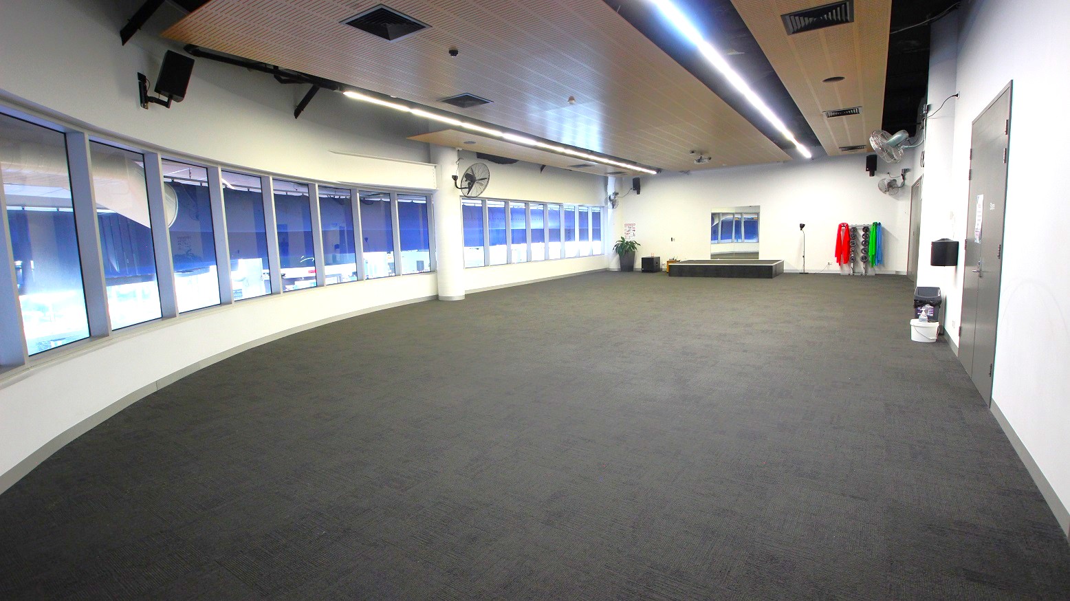 A spacious room with a curved wall of large windows on the left, allowing natural light to enter. The floor is covered with grey carpet. On the right, there is a wall with a door and some cleaning supplies. At the far end, there are exercise mats in various colours stacked vertically, a small platform, and a potted plant. The ceiling features recessed lighting and ventilation ducts. A spacious room with a curved wall of large windows on the left, allowing natural light to enter. The floor is covered with grey carpet. On the right, there is a wall with a door and some cleaning supplies. At the far end, there are exercise mats in various colours stacked vertically, a small platform, and a potted plant. The ceiling features recessed lighting and ventilation ducts.
