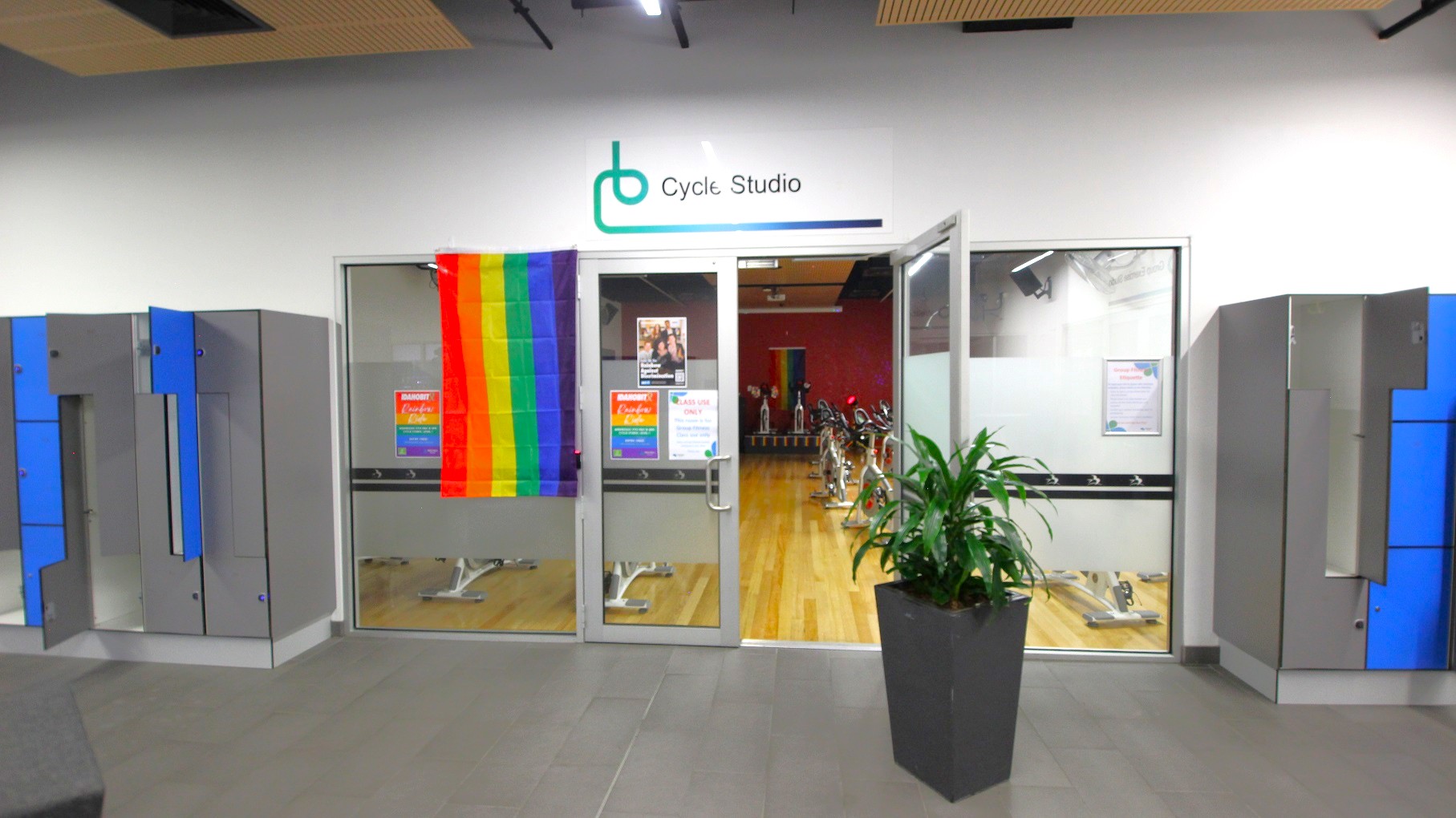 A cycle studio entrance with glass doors, displaying a rainbow flag on the left door. Above the doors, a sign reads "Cycle Studio". Inside, several stationary bikes are visible on a wooden floor. On the left and right of the entrance, there are grey lockers with blue doors, some of which are open. A large potted plant is positioned to the right of the entrance. The ceiling has wooden panels, and the floor is grey. A cycle studio entrance with glass doors, displaying a rainbow flag on the left door. Above the doors, a sign reads "Cycle Studio". Inside, several stationary bikes are visible on a wooden floor. On the left and right of the entrance, there are grey lockers with blue doors, some of which are open. A large potted plant is positioned to the right of the entrance. The ceiling has wooden panels, and the floor is grey.