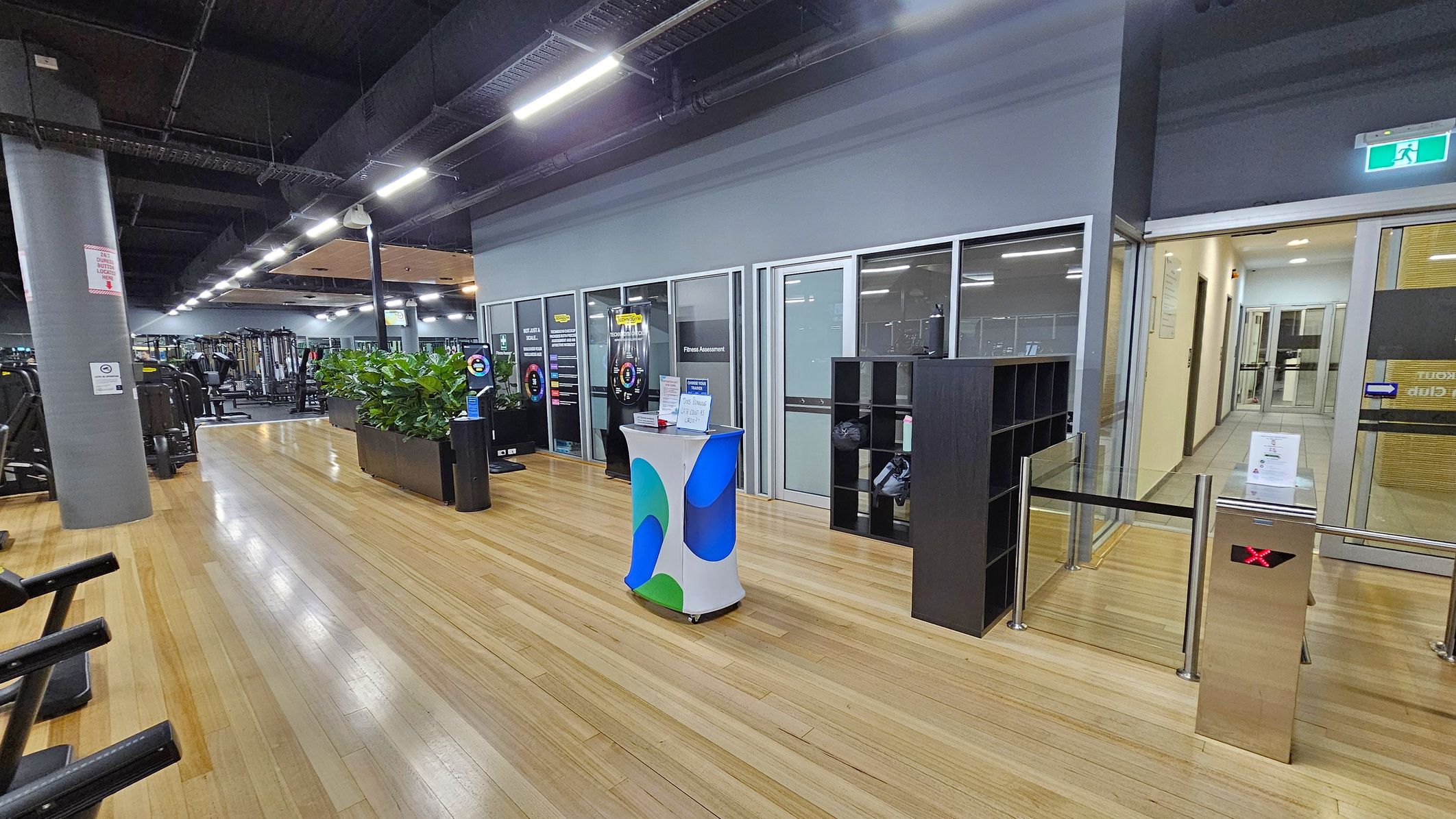 A gym interior with a wooden floor and a dark ceiling. On the left, there are exercise machines and a large grey pillar. In the centre, a small table with a colourful cover is placed near a row of glass-walled rooms labelled for fitness assessments. A black shelving unit is nearby. On the right, a turnstile with a red 'X' and a glass door leading to a hallway. Bright overhead lights illuminate the space, and a green exit sign is visible above the door. A gym interior with a wooden floor and a dark ceiling. On the left, there are exercise machines and a large grey pillar. In the centre, a small table with a colourful cover is placed near a row of glass-walled rooms labelled for fitness assessments. A black shelving unit is nearby. On the right, a turnstile with a red 'X' and a glass door leading to a hallway. Bright overhead lights illuminate the space, and a green exit sign is visible above the door.