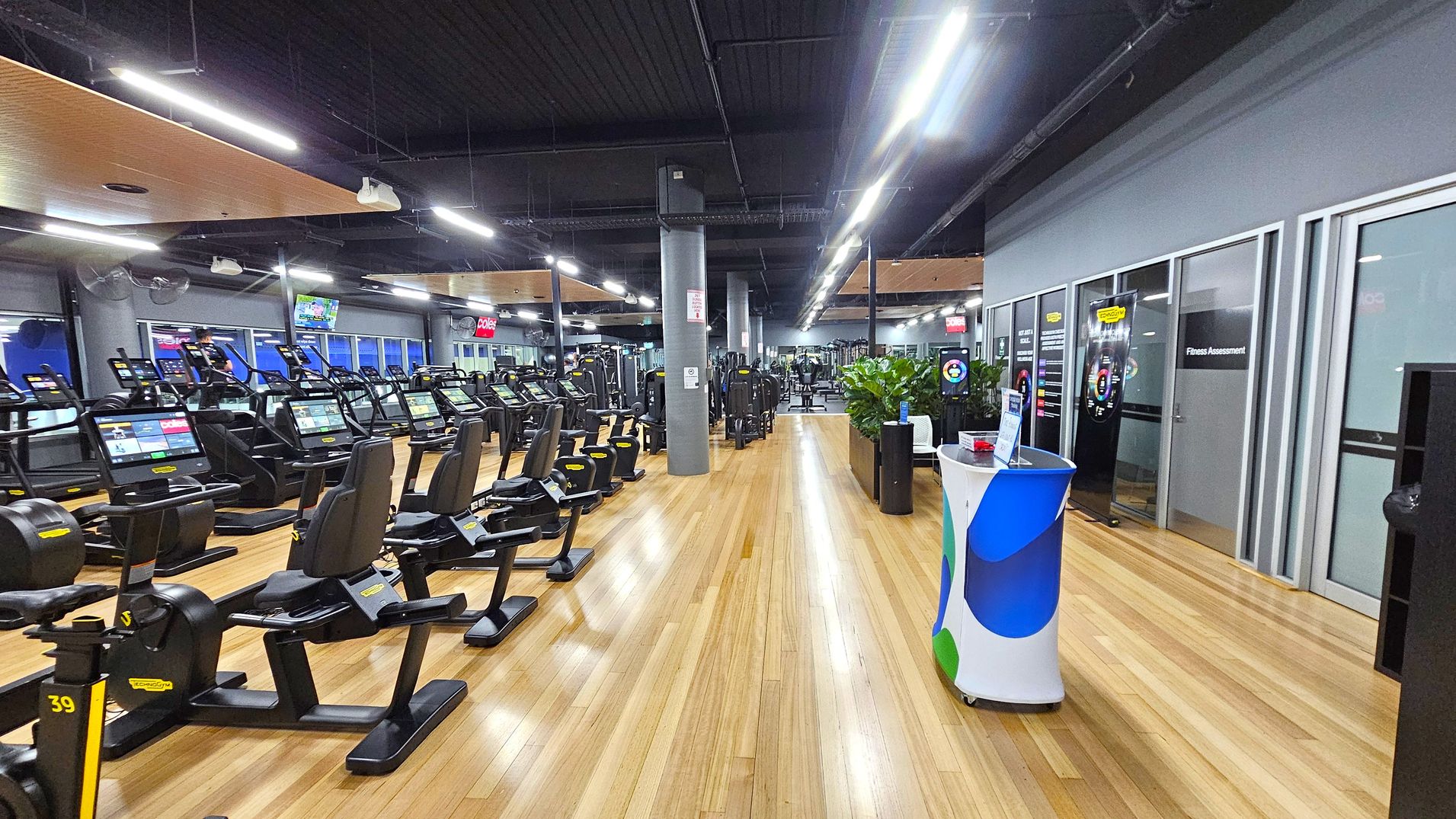 A spacious gym interior with a wooden floor and a dark ceiling. On the left, there are rows of exercise machines, including treadmills and stationary bikes, each equipped with digital screens. In the middle, a wide walkway leads to more equipment in the background. On the right, there are glass doors labelled "Fitness Assessment" and a small counter with a colourful cover. Potted plants are placed near the counter, adding greenery to the space. Bright overhead lights illuminate the area. A spacious gym interior with a wooden floor and a dark ceiling. On the left, there are rows of exercise machines, including treadmills and stationary bikes, each equipped with digital screens. In the middle, a wide walkway leads to more equipment in the background. On the right, there are glass doors labelled "Fitness Assessment" and a small counter with a colourful cover. Potted plants are placed near the counter, adding greenery to the space. Bright overhead lights illuminate the area.