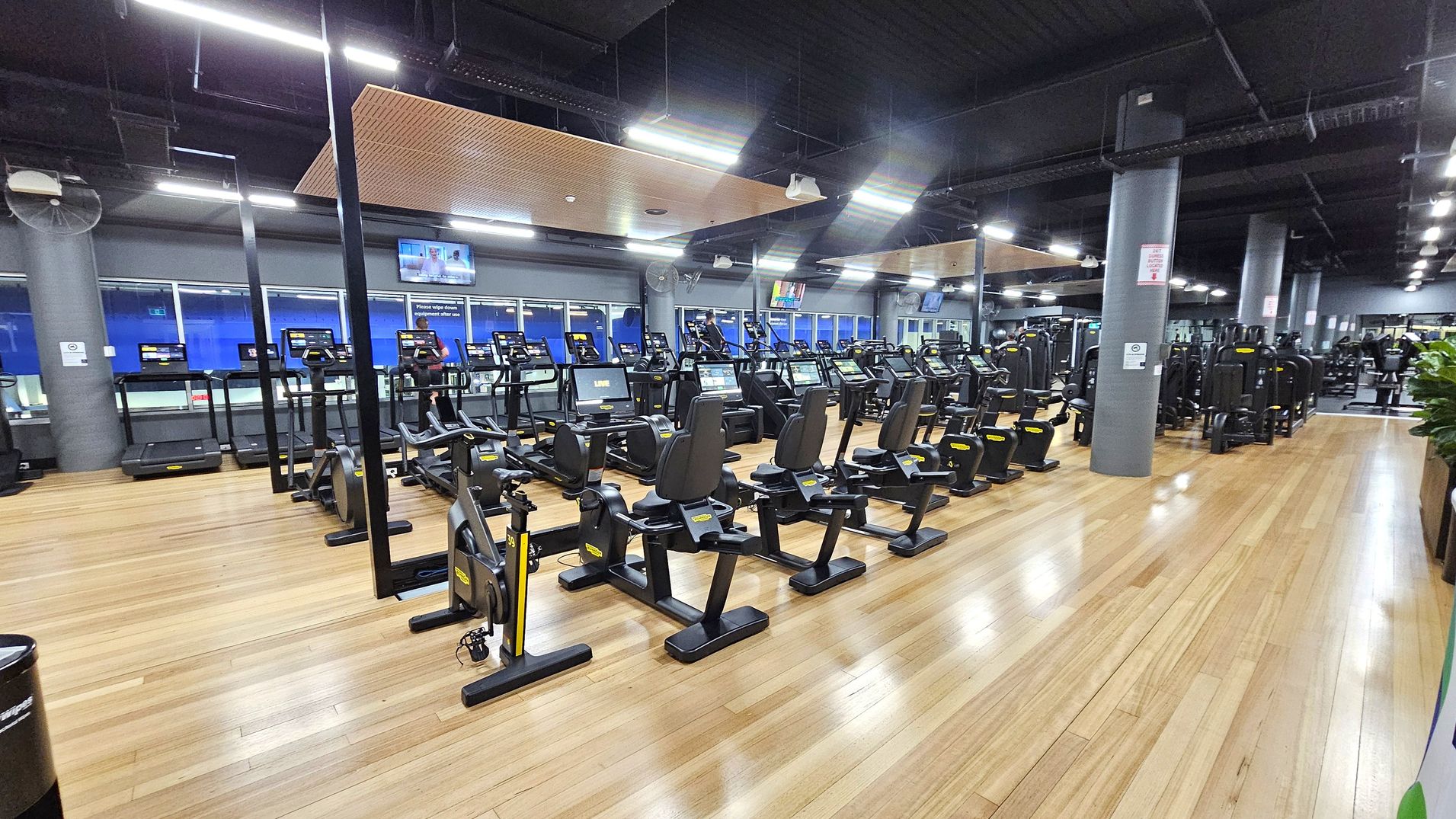 A spacious gym with wooden flooring and a dark ceiling. In the foreground, there are several stationary bikes and treadmills arranged in rows. The background features large windows and multiple television screens mounted on the walls. The gym is well-lit with overhead lights, and there are grey pillars throughout the space. On the right, there are additional exercise machines and a row of plants. A spacious gym with wooden flooring and a dark ceiling. In the foreground, there are several stationary bikes and treadmills arranged in rows. The background features large windows and multiple television screens mounted on the walls. The gym is well-lit with overhead lights, and there are grey pillars throughout the space. On the right, there are additional exercise machines and a row of plants.