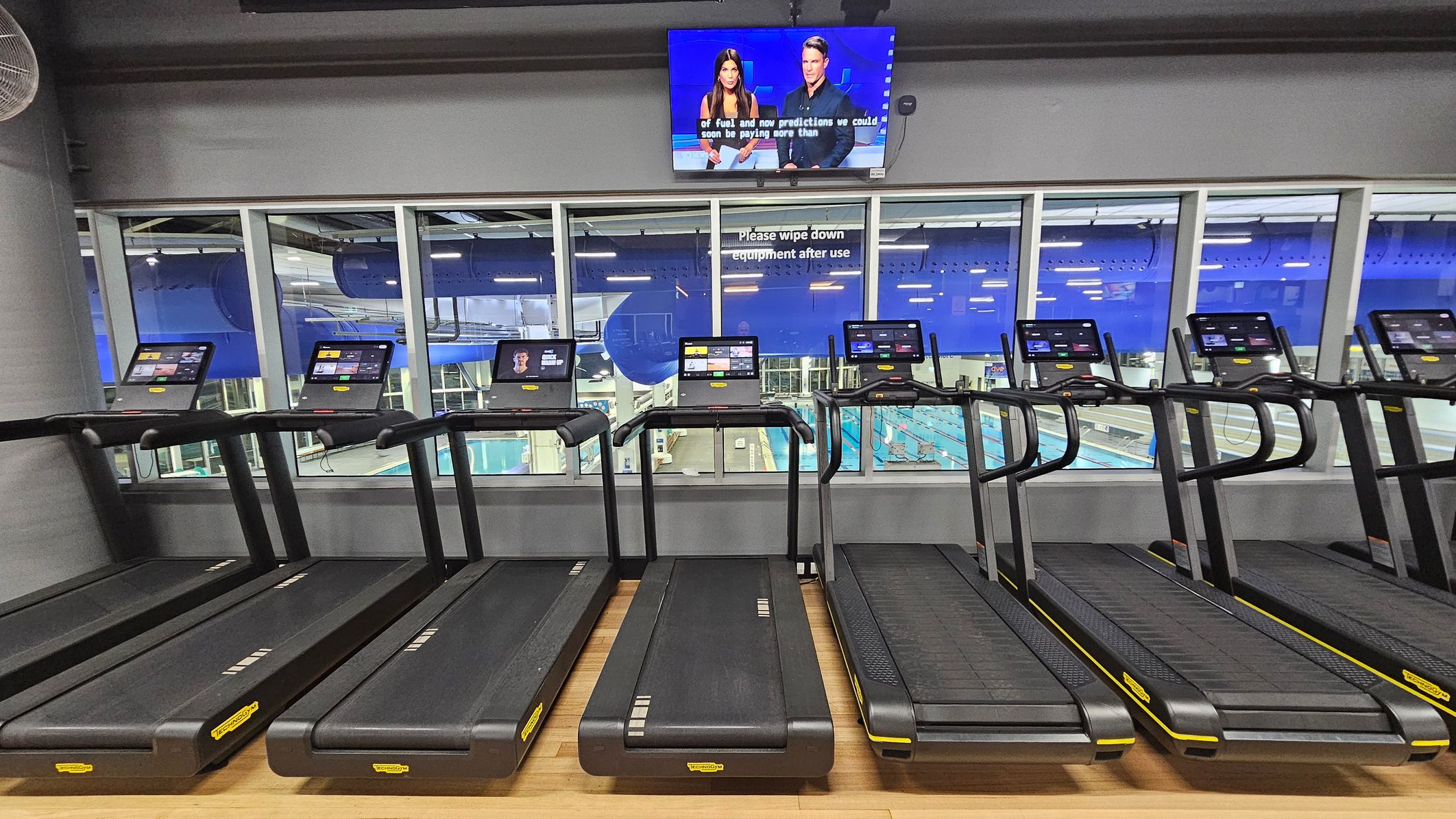 A row of treadmills with digital screens is positioned in the foreground on a wooden floor. Behind them, large windows reveal a view of an indoor swimming pool area with blue accents. Above the windows, a television screen displays a news broadcast featuring two presenters. A sign on the window reads, "Please wipe down equipment after use." The ceiling is industrial with visible ducts and lighting. A row of treadmills with digital screens is positioned in the foreground on a wooden floor. Behind them, large windows reveal a view of an indoor swimming pool area with blue accents. Above the windows, a television screen displays a news broadcast featuring two presenters. A sign on the window reads, "Please wipe down equipment after use." The ceiling is industrial with visible ducts and lighting.
