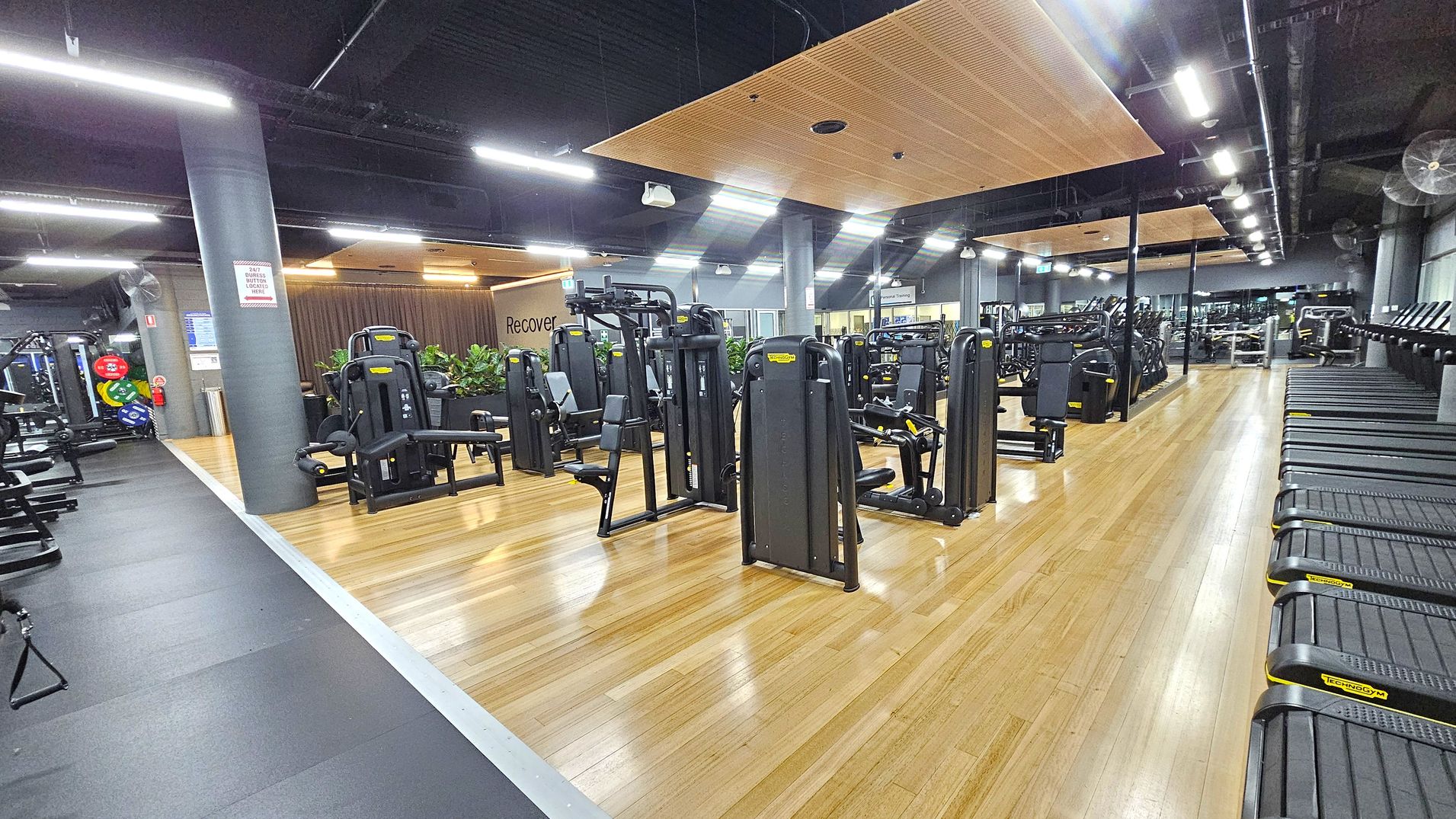 A spacious gym interior with wooden flooring. On the left, there are various weight machines and a column with a safety sign. In the centre, more exercise machines are arranged neatly, with a "Recovery" sign on the wall behind them. On the right, a row of treadmills lines the wall. The ceiling is dark with exposed beams and bright overhead lights. A spacious gym interior with wooden flooring. On the left, there are various weight machines and a column with a safety sign. In the centre, more exercise machines are arranged neatly, with a "Recovery" sign on the wall behind them. On the right, a row of treadmills lines the wall. The ceiling is dark with exposed beams and bright overhead lights.