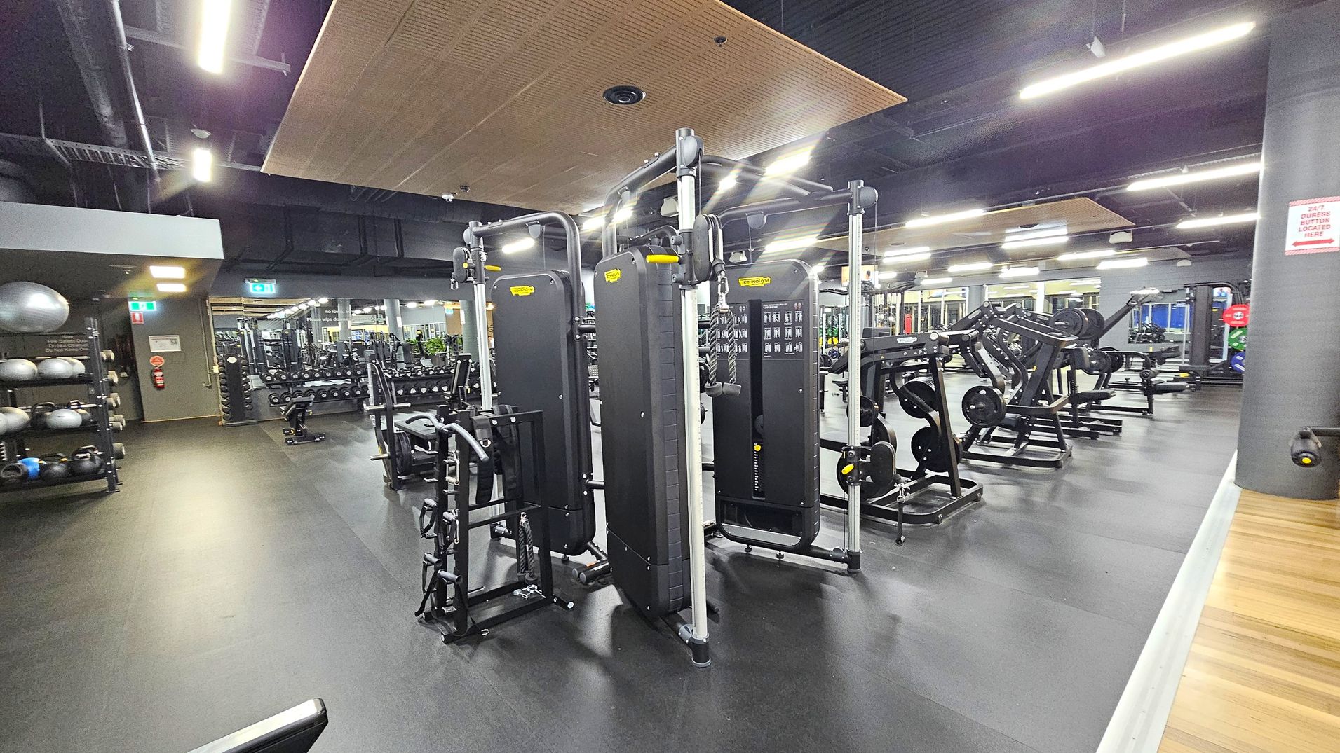 A spacious gym interior with a variety of exercise equipment. On the left, there are shelves with medicine balls and kettlebells. In the centre, several cable machines are arranged in a row. To the right, there are weight machines and benches. The floor is dark and rubberised, while the ceiling features exposed ducts and wooden panels. Bright overhead lights illuminate the space, and mirrors line the walls, reflecting the equipment. A spacious gym interior with a variety of exercise equipment. On the left, there are shelves with medicine balls and kettlebells. In the centre, several cable machines are arranged in a row. To the right, there are weight machines and benches. The floor is dark and rubberised, while the ceiling features exposed ducts and wooden panels. Bright overhead lights illuminate the space, and mirrors line the walls, reflecting the equipment.