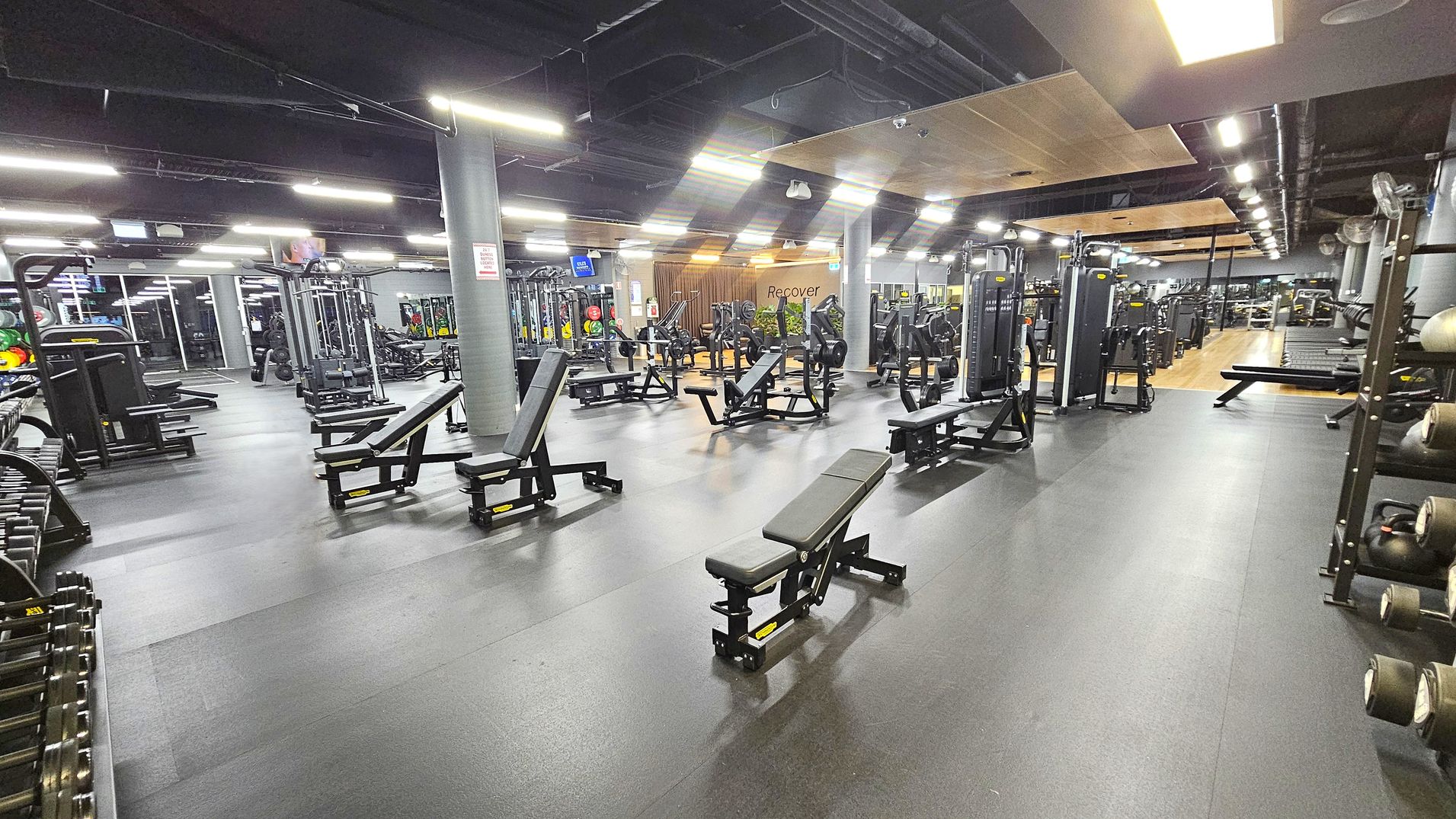 A spacious gym with various workout equipment. On the left, there are weight racks and benches. In the centre, multiple benches and weight machines are arranged neatly. On the right, more machines and a rack with medicine balls are visible. The floor is dark and smooth, while the ceiling has exposed ducts and bright lighting. In the background, a wooden panelled area with the word "Recover" is visible, along with more equipment and mirrors. A spacious gym with various workout equipment. On the left, there are weight racks and benches. In the centre, multiple benches and weight machines are arranged neatly. On the right, more machines and a rack with medicine balls are visible. The floor is dark and smooth, while the ceiling has exposed ducts and bright lighting. In the background, a wooden panelled area with the word "Recover" is visible, along with more equipment and mirrors.