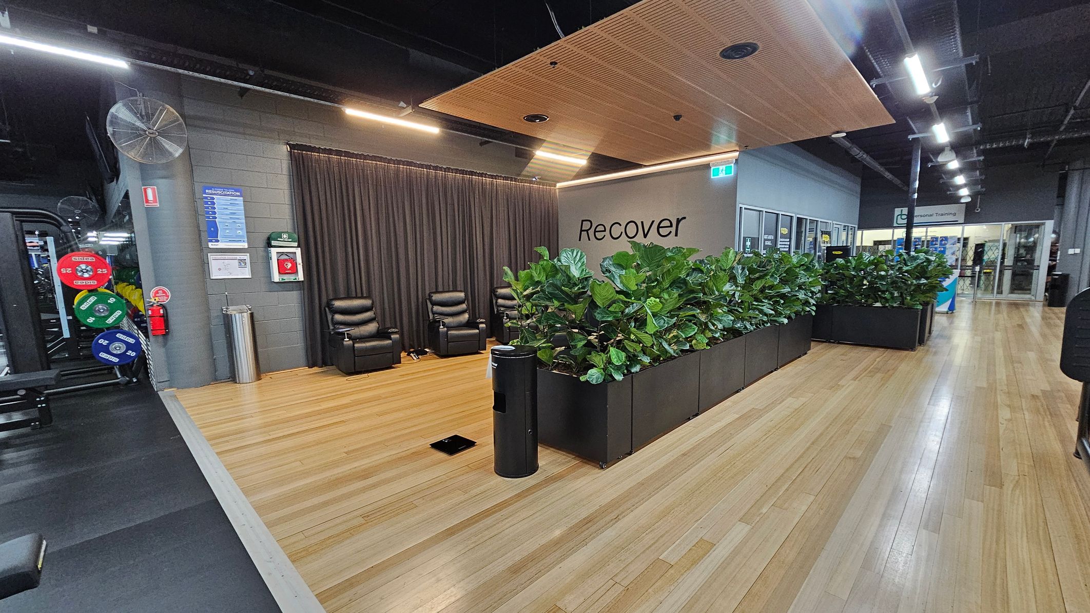 A gym recovery area with wooden flooring. On the left, there are weight plates and gym equipment. In the centre, two black recliner chairs are positioned against a dark curtain. A wall with the word "Recover" is visible, with a row of green plants in black planters in front. On the right, a corridor leads to a glass entrance with signs for personal training. The ceiling has wooden panels and exposed lighting fixtures. A gym recovery area with wooden flooring. On the left, there are weight plates and gym equipment. In the centre, two black recliner chairs are positioned against a dark curtain. A wall with the word "Recover" is visible, with a row of green plants in black planters in front. On the right, a corridor leads to a glass entrance with signs for personal training. The ceiling has wooden panels and exposed lighting fixtures.