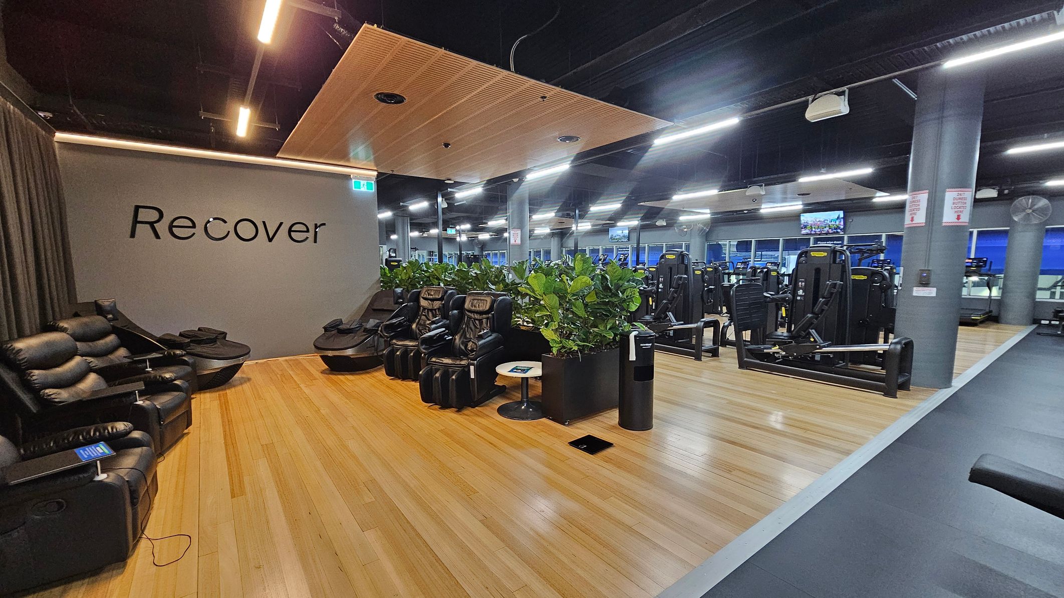 A gym interior with a recovery area on the left featuring black massage chairs and a grey wall with the word "Recover". The floor is wooden, and a small round table is nearby. In the centre, there are potted plants separating the recovery area from the gym equipment. On the right, various exercise machines are visible. The ceiling is dark with exposed fixtures and bright linear lights. A gym interior with a recovery area on the left featuring black massage chairs and a grey wall with the word "Recover". The floor is wooden, and a small round table is nearby. In the centre, there are potted plants separating the recovery area from the gym equipment. On the right, various exercise machines are visible. The ceiling is dark with exposed fixtures and bright linear lights.
