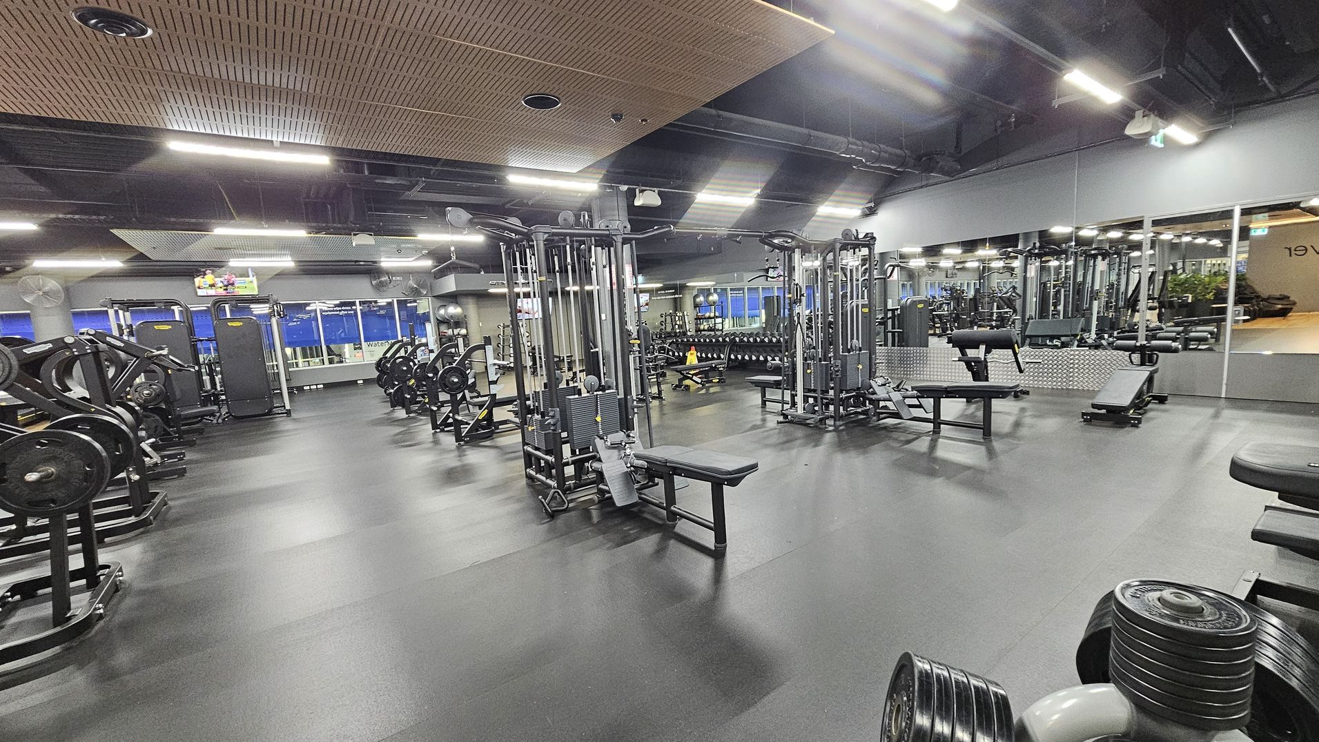 A spacious gym with a variety of exercise equipment. On the left, there are weight machines and benches. In the centre, there are cable machines and benches. The right side features a mirrored wall reflecting the gym equipment and a row of dumbbells. The floor is dark and rubberised, while the ceiling has a mix of exposed ducts and wooden panels. Bright overhead lights illuminate the space, and a TV is mounted on the wall in the background. A spacious gym with a variety of exercise equipment. On the left, there are weight machines and benches. In the centre, there are cable machines and benches. The right side features a mirrored wall reflecting the gym equipment and a row of dumbbells. The floor is dark and rubberised, while the ceiling has a mix of exposed ducts and wooden panels. Bright overhead lights illuminate the space, and a TV is mounted on the wall in the background.