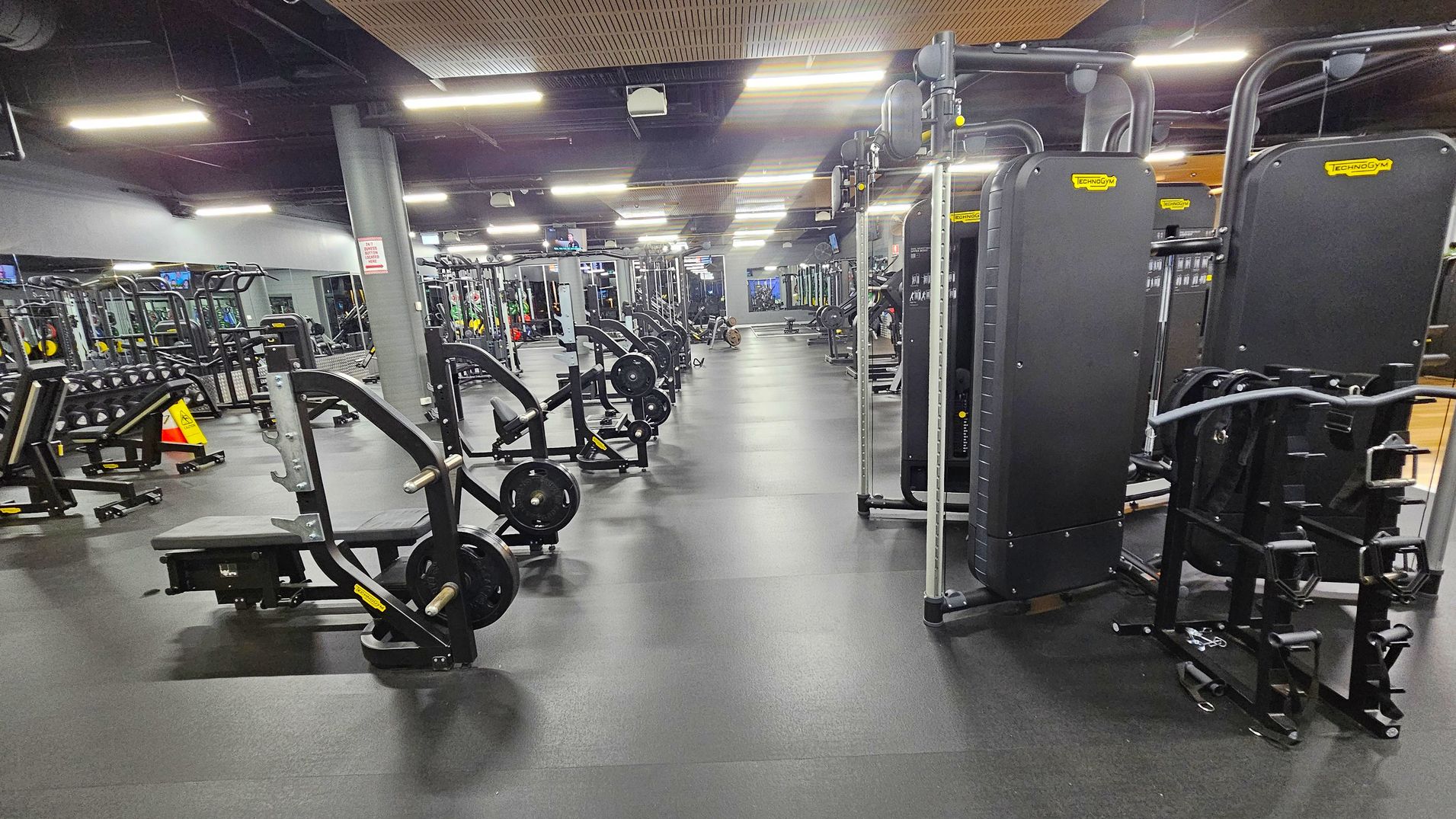 A spacious gym with various exercise machines. On the left, there are weight benches and racks with dumbbells. In the centre, multiple weightlifting machines are aligned in rows. On the right, there are large, upright fitness machines. The floor is covered with dark rubber mats, and the ceiling has exposed pipes and bright fluorescent lights. A grey pillar stands near the centre, and a caution sign is visible on the left. A spacious gym with various exercise machines. On the left, there are weight benches and racks with dumbbells. In the centre, multiple weightlifting machines are aligned in rows. On the right, there are large, upright fitness machines. The floor is covered with dark rubber mats, and the ceiling has exposed pipes and bright fluorescent lights. A grey pillar stands near the centre, and a caution sign is visible on the left.