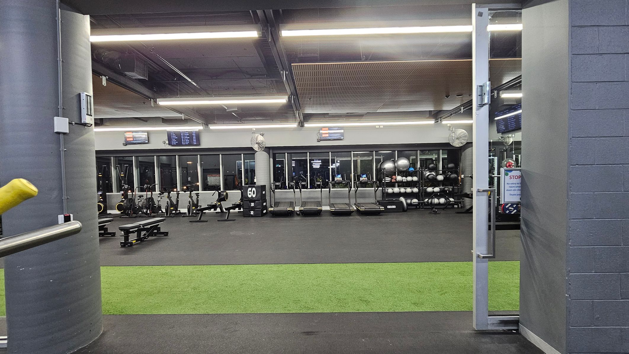 A gym interior with a grey and green colour scheme. In the foreground, a green artificial turf strip is visible on the floor. The background features a row of treadmills and exercise bikes against a wall with large windows. To the right, shelves hold exercise balls and other equipment. The ceiling has exposed beams and fluorescent lighting. A grey pillar is on the left, and a partially open door is on the right. A gym interior with a grey and green colour scheme. In the foreground, a green artificial turf strip is visible on the floor. The background features a row of treadmills and exercise bikes against a wall with large windows. To the right, shelves hold exercise balls and other equipment. The ceiling has exposed beams and fluorescent lighting. A grey pillar is on the left, and a partially open door is on the right.