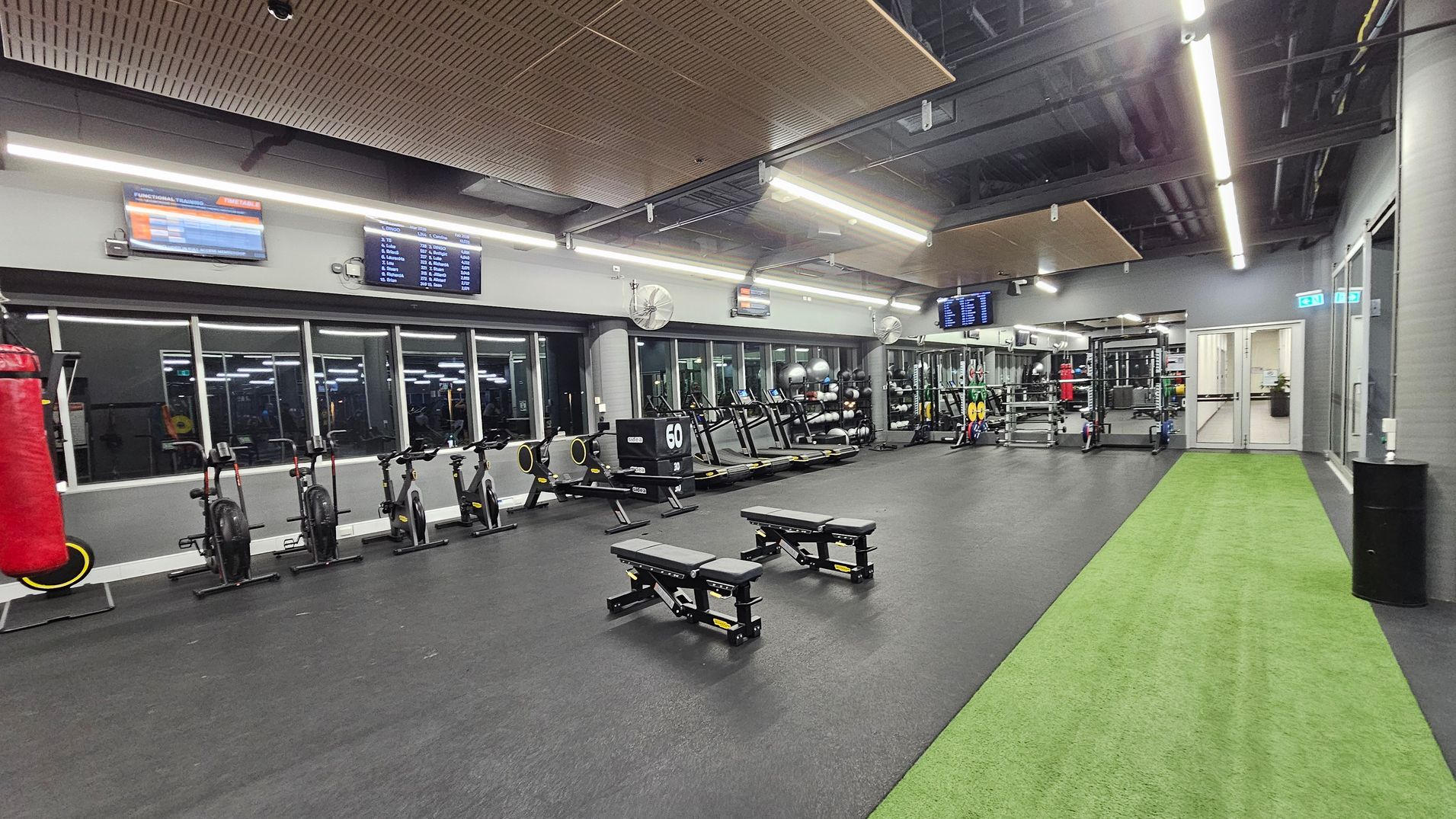 A spacious gym interior with a black rubber floor and a green artificial turf strip on the right. On the left, there are stationary bikes and a red punching bag. In the centre, two adjustable benches are positioned. The back wall features large mirrors and windows, with various gym equipment like treadmills and weights. The ceiling has exposed ducts and wooden panels, with bright fluorescent lighting. Several screens displaying information are mounted on the walls. A spacious gym interior with a black rubber floor and a green artificial turf strip on the right. On the left, there are stationary bikes and a red punching bag. In the centre, two adjustable benches are positioned. The back wall features large mirrors and windows, with various gym equipment like treadmills and weights. The ceiling has exposed ducts and wooden panels, with bright fluorescent lighting. Several screens displaying information are mounted on the walls.