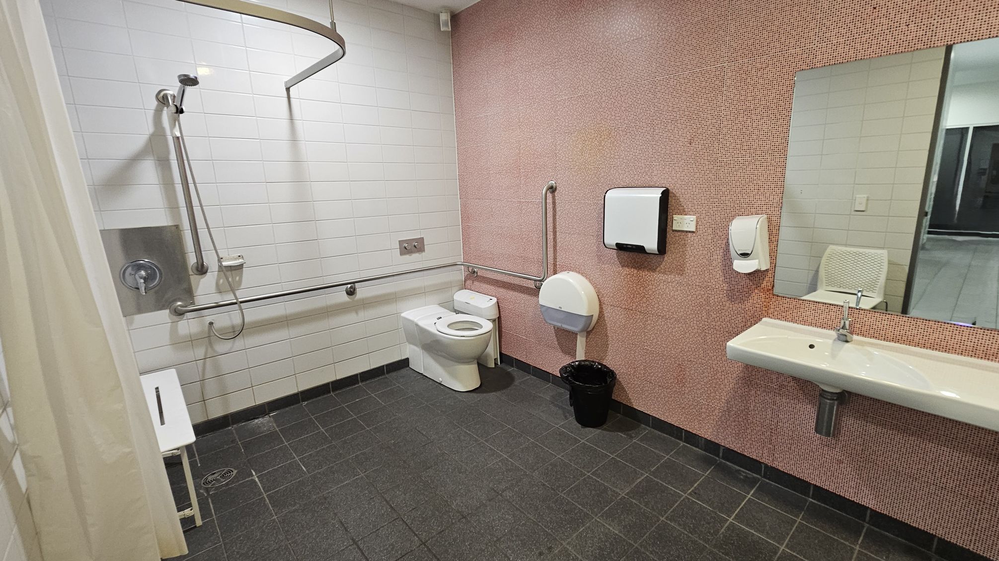 A spacious accessible bathroom with a dark tiled floor. On the left, there is a shower area with a handheld showerhead, grab bars, and a white shower seat. The back wall has white tiles. In the centre, a toilet with grab bars on both sides is visible. On the right, a pink-tiled wall features a large mirror above a white sink. A paper towel dispenser and soap dispenser are mounted on the wall. A black waste bin is on the floor. The ceiling is plain white. A spacious accessible bathroom with a dark tiled floor. On the left, there is a shower area with a handheld showerhead, grab bars, and a white shower seat. The back wall has white tiles. In the centre, a toilet with grab bars on both sides is visible. On the right, a pink-tiled wall features a large mirror above a white sink. A paper towel dispenser and soap dispenser are mounted on the wall. A black waste bin is on the floor. The ceiling is plain white.