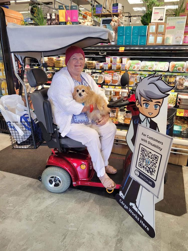 An elderly person in a scooter holds a small dog beside a Cérge cartoon cutout at a store.