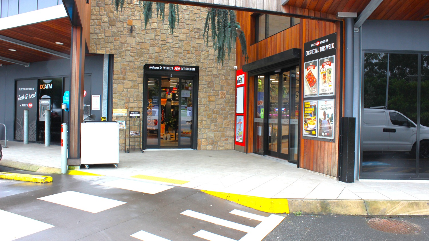 To the left, there are grey poles and a cash machine marked as "DCATM" next to a small shopping cart corral. The path leads to a slightly elevated concrete area where a white storage unit is positioned. On the right side, two large glass doors open into a store labelled "WHITES MT COOLOM", with promotional signs displayed next to them. The floor is a mixture of concrete and tiles with yellow and white lines marking pedestrian crossings. To the left, there are grey poles and a cash machine marked as "DCATM" next to a small shopping cart corral. The path leads to a slightly elevated concrete area where a white storage unit is positioned. On the right side, two large glass doors open into a store labelled "WHITES MT COOLOM", with promotional signs displayed next to them. The floor is a mixture of concrete and tiles with yellow and white lines marking pedestrian crossings.