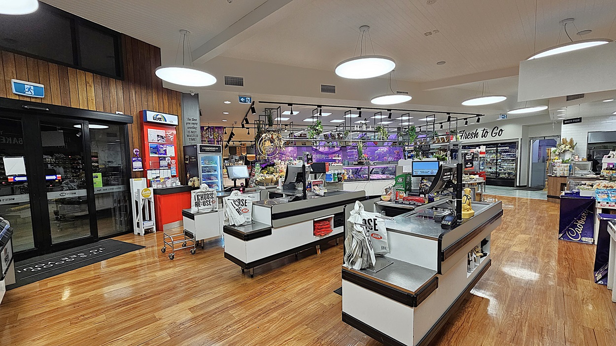 A supermarket with polished wooden floors. On the left, there is a glass entrance door, a red ATM, and a small display of magazines and brochures. In the middle, there are two checkout counters with black and white designs, each with a conveyor belt and shopping bags. The ceiling has white panels with large circular lights. On the right, there are shelves with flowers, a "Flowers To Go" sign, and a display of Cadbury Dairy Milk chocolates. A supermarket with polished wooden floors. On the left, there is a glass entrance door, a red ATM, and a small display of magazines and brochures. In the middle, there are two checkout counters with black and white designs, each with a conveyor belt and shopping bags. The ceiling has white panels with large circular lights. On the right, there are shelves with flowers, a "Flowers To Go" sign, and a display of Cadbury Dairy Milk chocolates.
