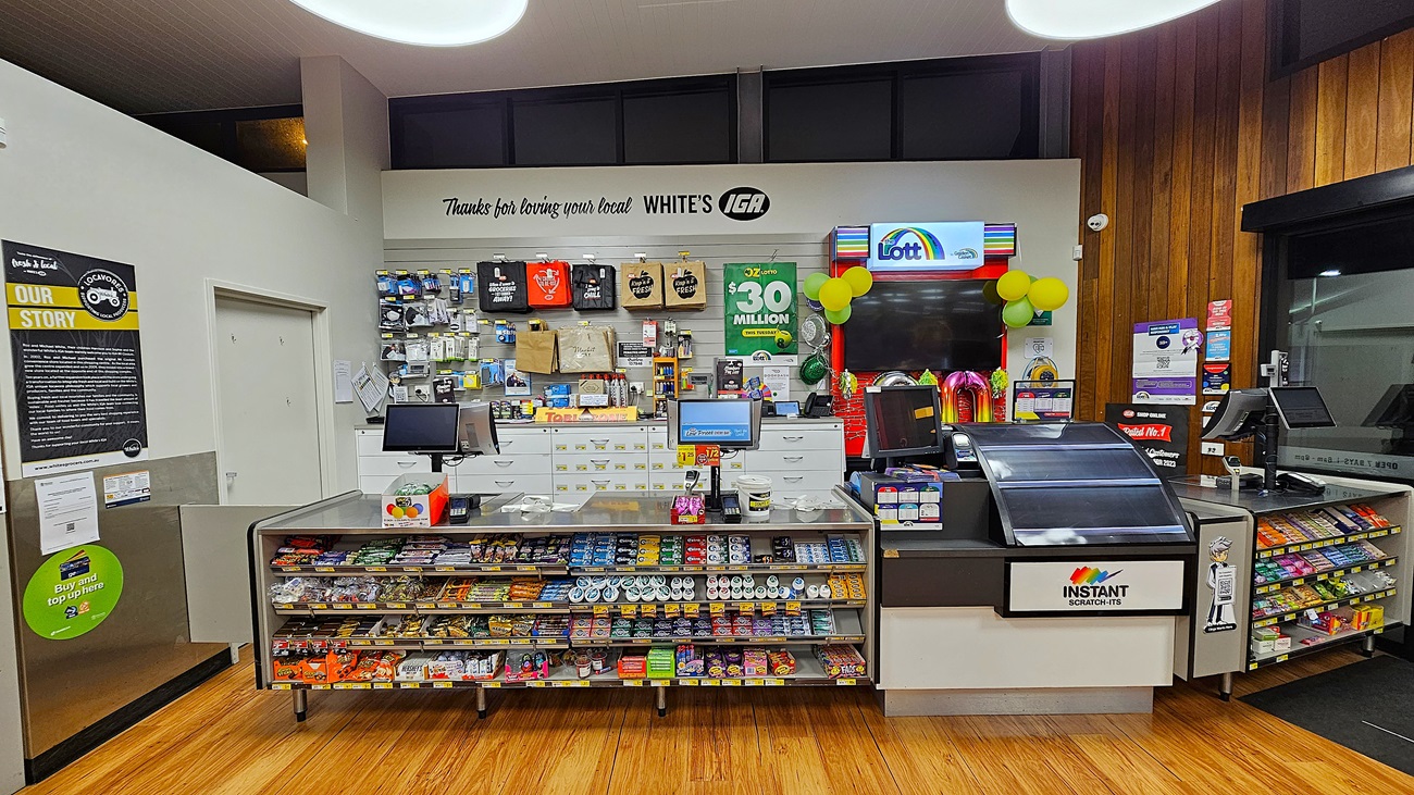 A supermarket with polished wooden floors. On the left, there is a white wall with a bulletin board and a door. In the middle, there are two checkout counters with various snacks, a display of gift cards, and a sign reading "Thanks for loving your local WHITE'S IGA." The ceiling has white panels with large circular lights. On the right, there is a lottery ticket machine with a colourful display and a window with a view outside. The background features more shelves with snacks and a "Lott" sign. A supermarket with polished wooden floors. On the left, there is a white wall with a bulletin board and a door. In the middle, there are two checkout counters with various snacks, a display of gift cards, and a sign reading "Thanks for loving your local WHITE'S IGA." The ceiling has white panels with large circular lights. On the right, there is a lottery ticket machine with a colourful display and a window with a view outside. The background features more shelves with snacks and a "Lott" sign.