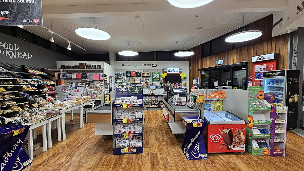 A supermarket with polished wooden floors. On the left, there are shelves with baked goods and a black wall with the words "FOOD YOU KNEAD." In the middle, there are two checkout counters with various snacks, a display of gift cards, and a sign reading "Thanks for loving your local WHITE'S IGA." The ceiling has white panels with large circular lights. On the right, there is a lottery ticket machine, a red ice cream freezer, and a window with a view outside. A supermarket with polished wooden floors. On the left, there are shelves with baked goods and a black wall with the words "FOOD YOU KNEAD." In the middle, there are two checkout counters with various snacks, a display of gift cards, and a sign reading "Thanks for loving your local WHITE'S IGA." The ceiling has white panels with large circular lights. On the right, there is a lottery ticket machine, a red ice cream freezer, and a window with a view outside.