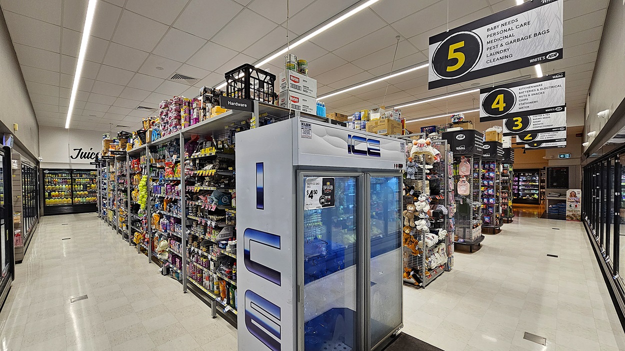 A supermarket aisle with a white tiled floor. On the left, there are refrigerated sections with glass doors displaying dairy products and juices. In the middle, there are shelves stocked with various household items, including light bulbs and batteries, and a large freezer labelled "ICE." The ceiling is white with rectangular fluorescent lights. On the right, there are more shelves with cleaning supplies and pet food, with overhead signs numbered 3 and 5 indicating different sections. A supermarket aisle with a white tiled floor. On the left, there are refrigerated sections with glass doors displaying dairy products and juices. In the middle, there are shelves stocked with various household items, including light bulbs and batteries, and a large freezer labelled "ICE." The ceiling is white with rectangular fluorescent lights. On the right, there are more shelves with cleaning supplies and pet food, with overhead signs numbered 3 and 5 indicating different sections.