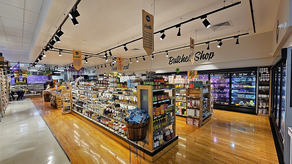 A supermarket with a mix of polished wooden and white tiled floors. On the left, there are shelves stocked with various packaged goods and a small wooden display table. In the middle, there is a large island with shelves filled with condiments, snacks, and a wooden barrel. The ceiling has white panels with black track lighting and hanging signs. On the right, there are refrigerated sections with glass doors displaying meats and dairy products, and a sign reading "Butcher Shop." A supermarket with a mix of polished wooden and white tiled floors. On the left, there are shelves stocked with various packaged goods and a small wooden display table. In the middle, there is a large island with shelves filled with condiments, snacks, and a wooden barrel. The ceiling has white panels with black track lighting and hanging signs. On the right, there are refrigerated sections with glass doors displaying meats and dairy products, and a sign reading "Butcher Shop."