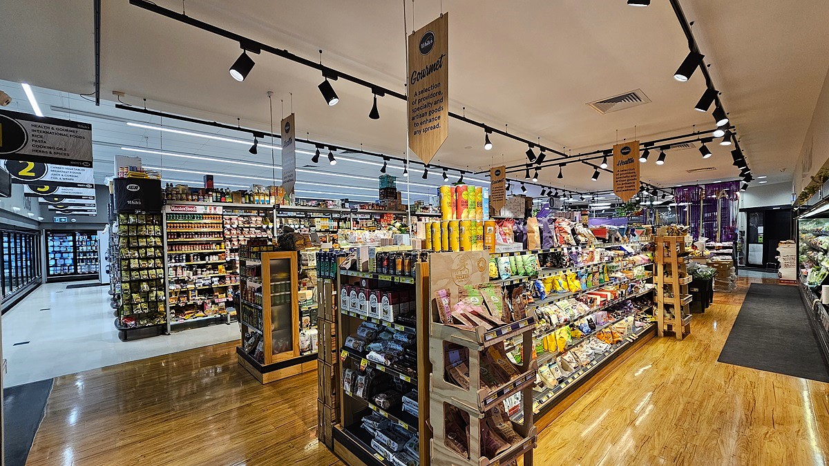 A supermarket with a mix of polished wooden and white tiled floors. On the left, there are shelves stocked with various packaged goods and a refrigerated section with glass doors displaying frozen items. In the middle, there are wooden display tables filled with snacks, condiments, and beverages, with hanging signs above. The ceiling has white panels with black track lighting. On the right, there are more shelves with fresh produce and a refrigerated section with dairy products. A supermarket with a mix of polished wooden and white tiled floors. On the left, there are shelves stocked with various packaged goods and a refrigerated section with glass doors displaying frozen items. In the middle, there are wooden display tables filled with snacks, condiments, and beverages, with hanging signs above. The ceiling has white panels with black track lighting. On the right, there are more shelves with fresh produce and a refrigerated section with dairy products.