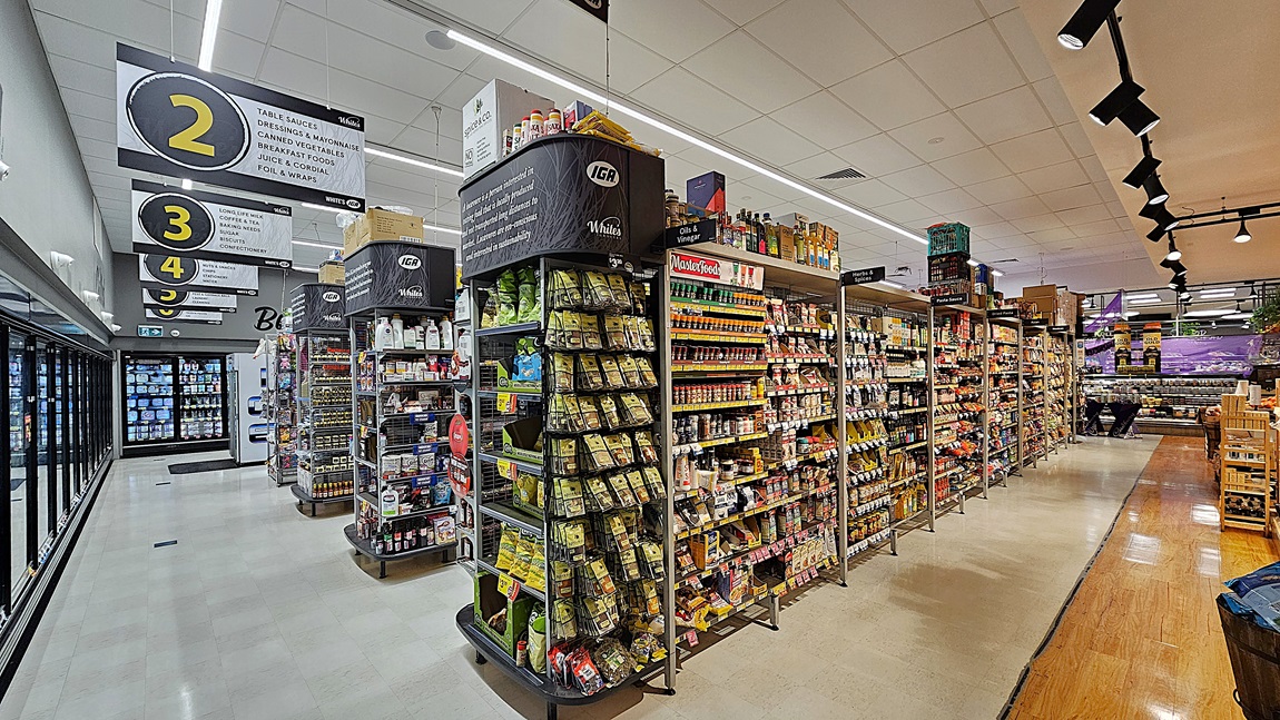 A supermarket aisle with a white tiled floor and a section of wooden flooring. On the left, there are tall shelves stocked with various snacks, including crisps and nuts, and a refrigerated section with glass doors displaying dairy products. In the middle, there are overhead signs numbered 2, 3, and 4 indicating different sections. The ceiling is white with rectangular fluorescent lights and black track lighting. On the right, there are more shelves filled with canned goods and condiments. A supermarket aisle with a white tiled floor and a section of wooden flooring. On the left, there are tall shelves stocked with various snacks, including crisps and nuts, and a refrigerated section with glass doors displaying dairy products. In the middle, there are overhead signs numbered 2, 3, and 4 indicating different sections. The ceiling is white with rectangular fluorescent lights and black track lighting. On the right, there are more shelves filled with canned goods and condiments.