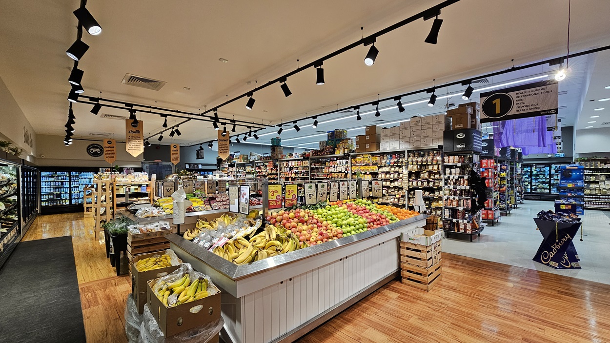 A supermarket with a mix of polished wooden and grey tiled floors. On the left, there are refrigerated sections with glass doors displaying beverages and dairy products. In the middle, there is a large display of fresh fruits, including bananas, apples, and oranges, with hanging signs above. The ceiling has white panels with black track lighting. On the right, there are shelves stocked with various packaged goods and a sign indicating aisle 1. A supermarket with a mix of polished wooden and grey tiled floors. On the left, there are refrigerated sections with glass doors displaying beverages and dairy products. In the middle, there is a large display of fresh fruits, including bananas, apples, and oranges, with hanging signs above. The ceiling has white panels with black track lighting. On the right, there are shelves stocked with various packaged goods and a sign indicating aisle 1.