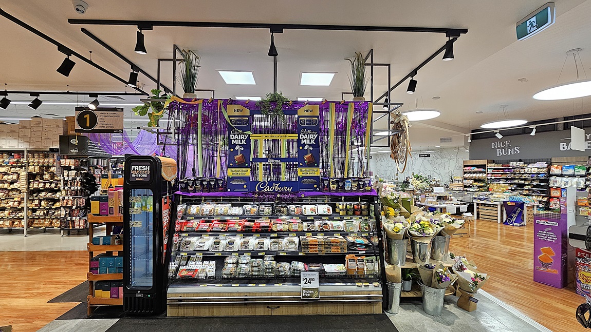 A supermarket with a mix of polished wooden and grey tiled floors. On the left, there is a black refrigerated unit with drinks and a sign indicating aisle 1. In the middle, there is a large display of Cadbury Dairy Milk chocolates, surrounded by purple decorations and hanging plants, with shelves of pre-packaged meals below. The ceiling has white panels with black track lighting. On the right, there are shelves with baked goods and a flower display in metal buckets. A supermarket with a mix of polished wooden and grey tiled floors. On the left, there is a black refrigerated unit with drinks and a sign indicating aisle 1. In the middle, there is a large display of Cadbury Dairy Milk chocolates, surrounded by purple decorations and hanging plants, with shelves of pre-packaged meals below. The ceiling has white panels with black track lighting. On the right, there are shelves with baked goods and a flower display in metal buckets.