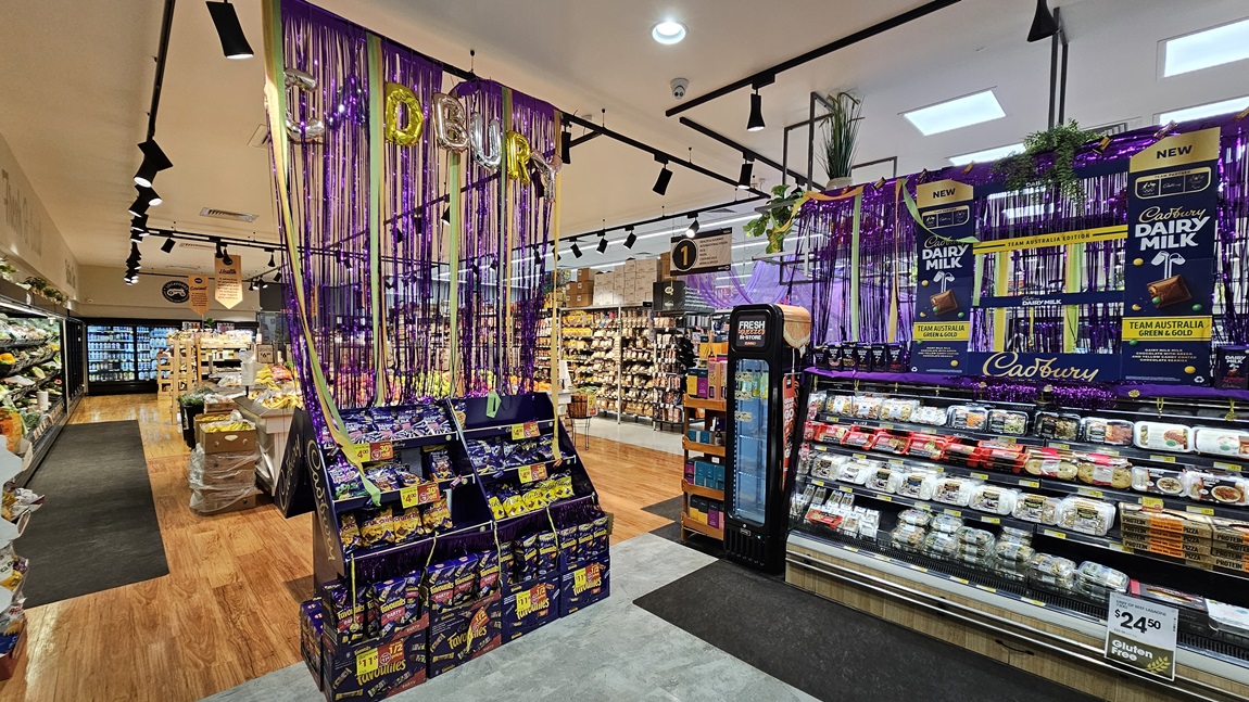 A supermarket with grey tiled and polished wooden floors. On the left, there is a large display of Cadbury Dairy Milk chocolates, decorated with purple streamers and hanging plants, with shelves of pre-packaged meals below. On the right, there are shelves with various food items, a fire extinguisher, and a sign reading "FOOD." The ceiling has white panels with black track lighting. In the background, there are more aisles, a flower display, and a sign that says "BAKE FOR MORE." A supermarket with grey tiled and polished wooden floors. On the left, there is a large display of Cadbury Dairy Milk chocolates, decorated with purple streamers and hanging plants, with shelves of pre-packaged meals below. On the right, there are shelves with various food items, a fire extinguisher, and a sign reading "FOOD." The ceiling has white panels with black track lighting. In the background, there are more aisles, a flower display, and a sign that says "BAKE FOR MORE."