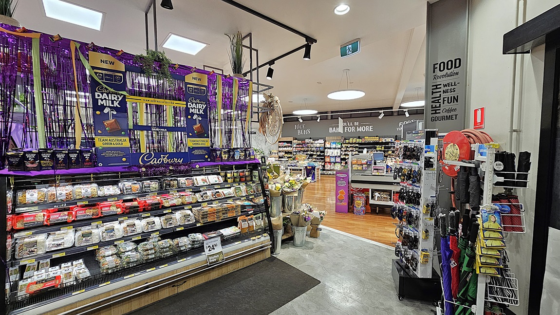 A supermarket with a mix of polished wooden and grey tiled floors. On the left, there are shelves with various packaged goods and a refrigerated section with glass doors displaying beverages. In the middle, there is a large display of Cadbury Dairy Milk chocolates, decorated with purple streamers and hanging plants. The ceiling has white panels with black track lighting. On the right, there are more Cadbury Dairy Milk products and a refrigerated section with pre-packaged meals. A supermarket with a mix of polished wooden and grey tiled floors. On the left, there are shelves with various packaged goods and a refrigerated section with glass doors displaying beverages. In the middle, there is a large display of Cadbury Dairy Milk chocolates, decorated with purple streamers and hanging plants. The ceiling has white panels with black track lighting. On the right, there are more Cadbury Dairy Milk products and a refrigerated section with pre-packaged meals.