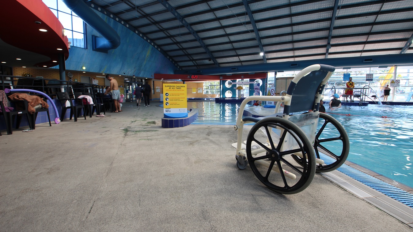 An aquatic wheelchair settled near the children’s pool. Black chairs are scattered around the pool for bags. At the end of the pools are glass walls that show the external pool area. An aquatic wheelchair settled near the children’s pool. Black chairs are scattered around the pool for bags. At the end of the pools are glass walls that show the external pool area.