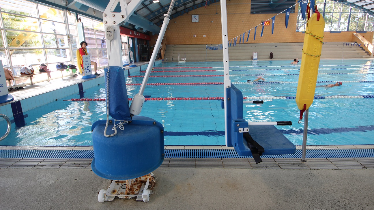 White pool hoist with blue seating chair at the 8-lane 25m lap pool. Near it is a yellow oblong floating platform used by lifeguards when saving someone. Long benches are placed against the glass walls for bags and other personal belongings. White pool hoist with blue seating chair at the 8-lane 25m lap pool. Near it is a yellow oblong floating platform used by lifeguards when saving someone. Long benches are placed against the glass walls for bags and other personal belongings.