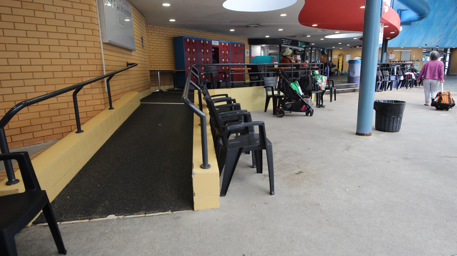 Entrance ramp with black smooth pathway, black handrails, and pale brown bricks leading to the red and blue locker cabinets and another seating area with mahogany table tops with metal frames and black chairs. A number of black chairs are designated slightly lower the ramp. The walls next to the Swell Café are made of bricks. Entrance ramp with black smooth pathway, black handrails, and pale brown bricks leading to the red and blue locker cabinets and another seating area with mahogany table tops with metal frames and black chairs. A number of black chairs are designated slightly lower the ramp. The walls next to the Swell Café are made of bricks.
