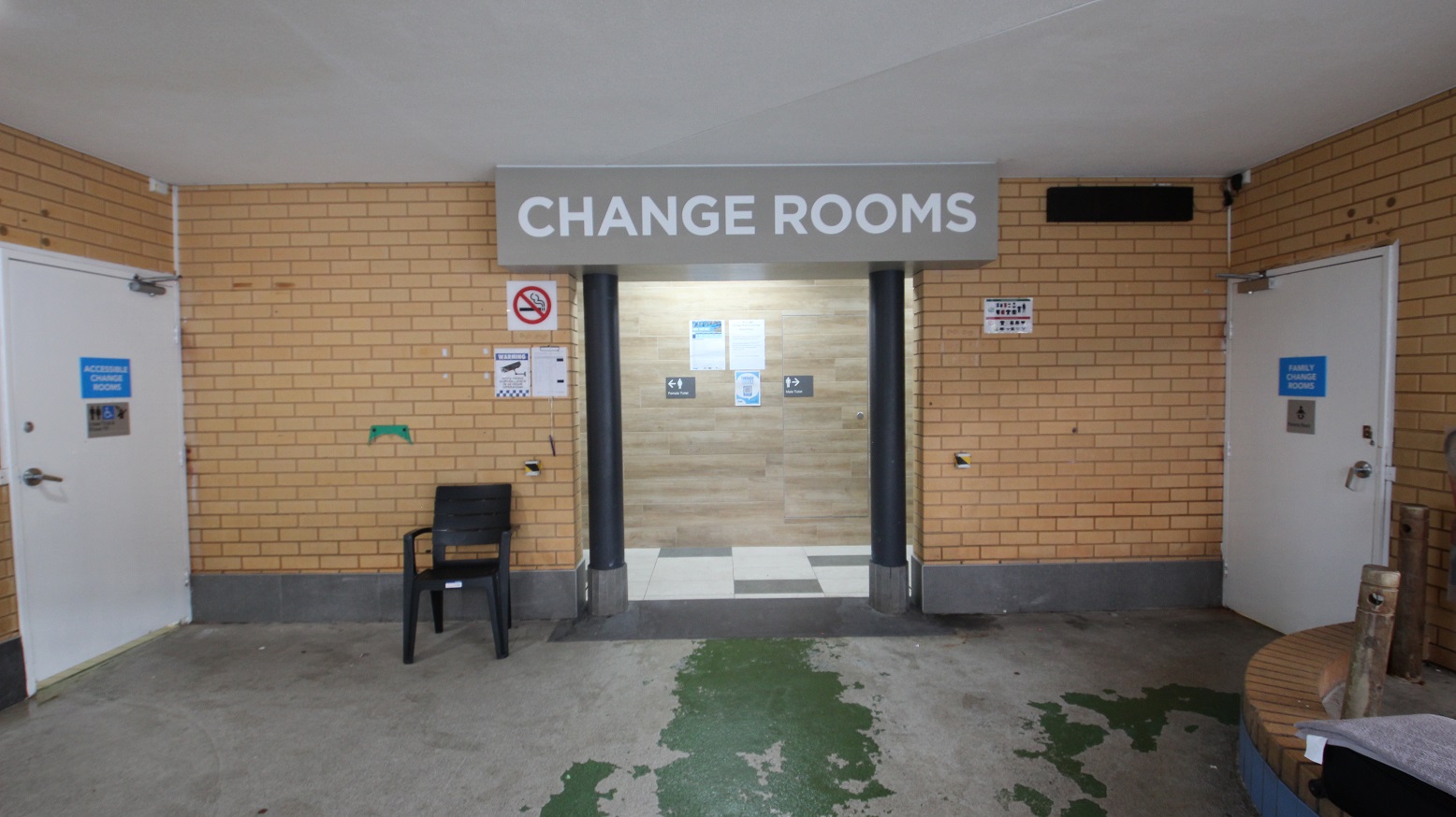 Change rooms at Whitlam Leisure Centre can be distinguished by the large brown block with black posts at the entrance. The ceiling is white, while the walls are made of bricks. Two white accessible change rooms opposite each other can be utilized by persons with mobility disabilities. A smoking sign and a CCTV sign are placed on the brick wall Change rooms at Whitlam Leisure Centre can be distinguished by the large brown block with black posts at the entrance. The ceiling is white, while the walls are made of bricks. Two white accessible change rooms opposite each other can be utilized by persons with mobility disabilities. A smoking sign and a CCTV sign are placed on the brick wall