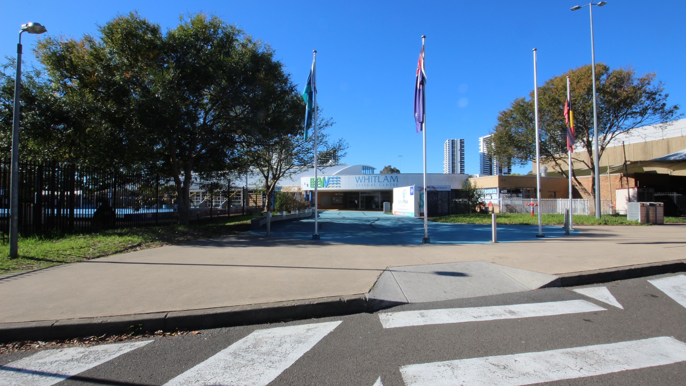 A zebra crossing leading to the smooth, concrete, blue pathway to the entrance of Whitlam Leisure Centre. At the boundary of the grey pathway are flag poles, flags, and grey bollards. There are also lamp posts assigned to opposite sides of this wide pathway. A large tree inside the black fence and pool provides great shade to the pathway. A zebra crossing leading to the smooth, concrete, blue pathway to the entrance of Whitlam Leisure Centre. At the boundary of the grey pathway are flag poles, flags, and grey bollards. There are also lamp posts assigned to opposite sides of this wide pathway. A large tree inside the black fence and pool provides great shade to the pathway.