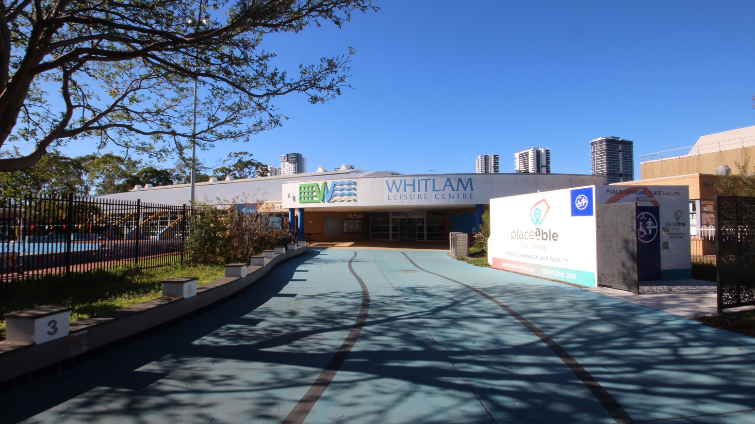 A smooth blue concrete pathway leading to Whitlam Leisure Centre. The venue is a low-set building with grey, white, and blue painting. The upper wall of the front face of the building shows the name of the venue and its logo. On the left is a 50-meter external pool secured by a black fence, while on the right is a public changing place facility by Placeable Solutions. The entry point to the facility is through a black gate. A smooth blue concrete pathway leading to Whitlam Leisure Centre. The venue is a low-set building with grey, white, and blue painting. The upper wall of the front face of the building shows the name of the venue and its logo. On the left is a 50-meter external pool secured by a black fence, while on the right is a public changing place facility by Placeable Solutions. The entry point to the facility is through a black gate.