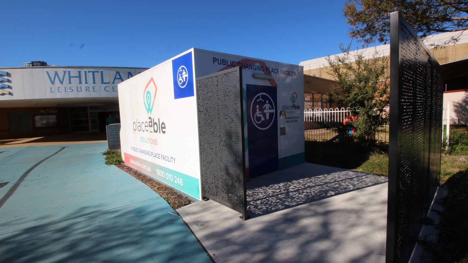 A closer look at the public changing place facility by Placeable Solutions. The right side of the black gate is opened and a square, dirty white, concrete pathway is placed in front of the facility. The facility has a dark blue door with accessible icons. A closer look at the public changing place facility by Placeable Solutions. The right side of the black gate is opened and a square, dirty white, concrete pathway is placed in front of the facility. The facility has a dark blue door with accessible icons.