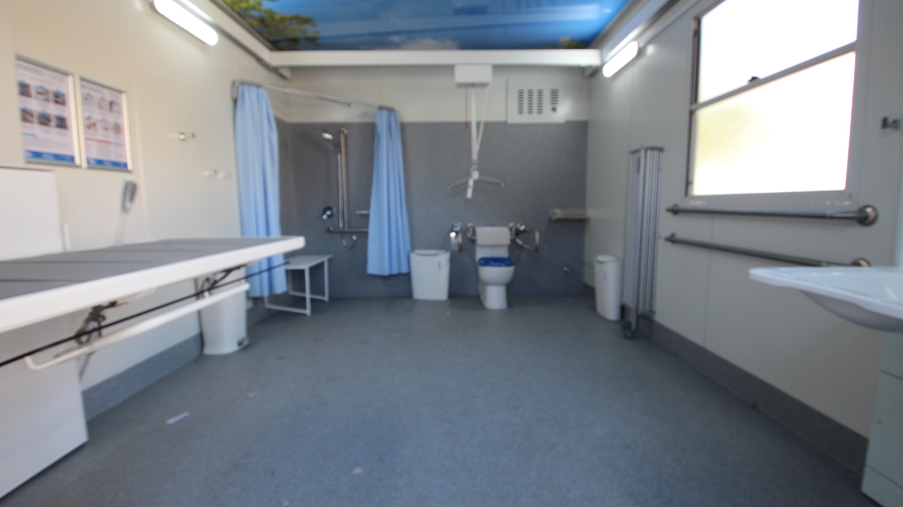 Inside the accessible bathroom at Whitlam Leisure Centre is a hoist, a dark blue cushioned adult change table, a toilet, and a shower with a sky blue privacy curtain. Beside the toilet bowl is a white rubbish bin. The back wall is painted grey, while the left and right are smooth white walls. Near the entrance, to the left, is a wash basin. Inside the accessible bathroom at Whitlam Leisure Centre is a hoist, a dark blue cushioned adult change table, a toilet, and a shower with a sky blue privacy curtain. Beside the toilet bowl is a white rubbish bin. The back wall is painted grey, while the left and right are smooth white walls. Near the entrance, to the left, is a wash basin.