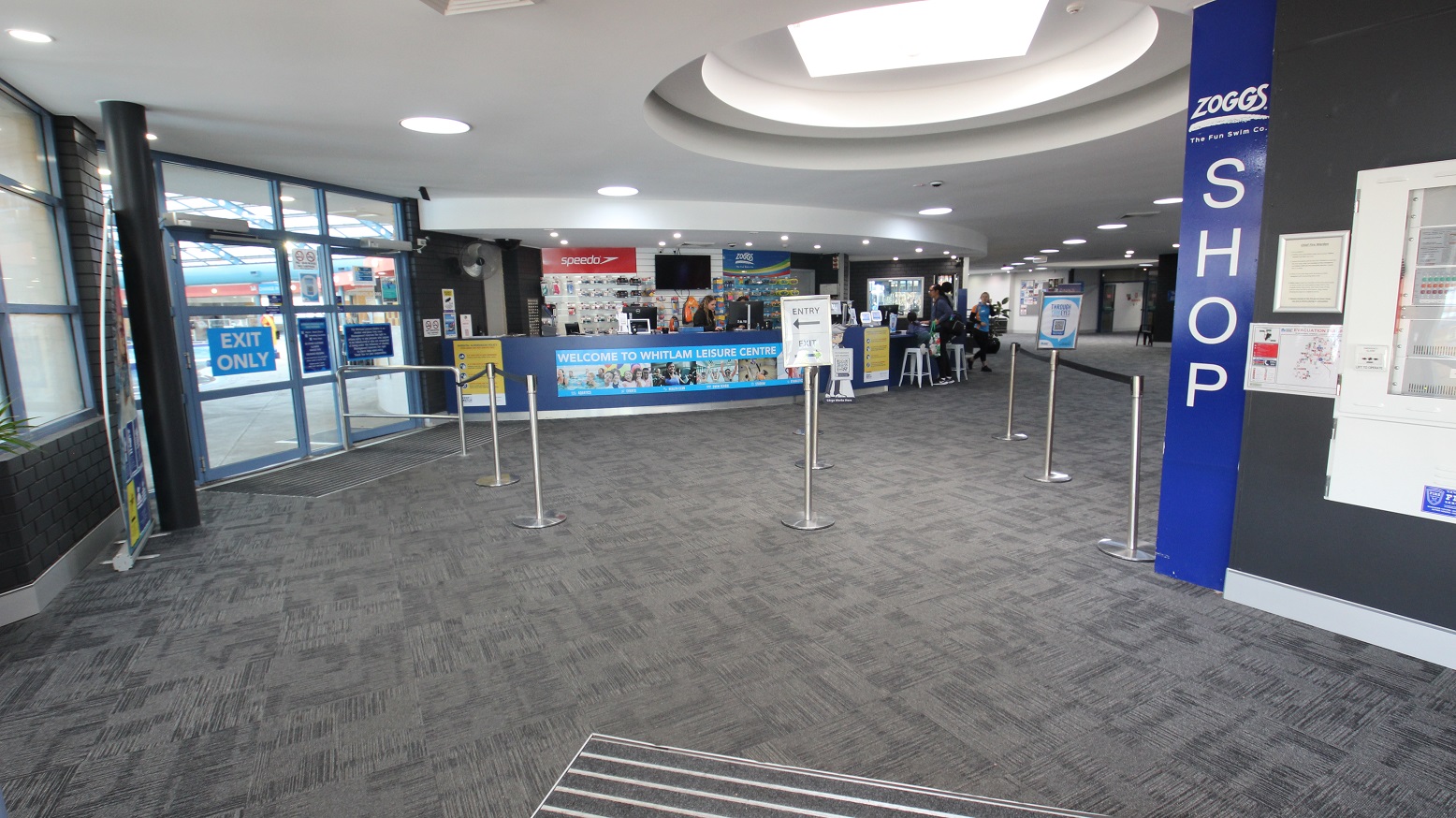 The image shows the view inside Whitlam Leisure Centre after passing through the entrance and exit doors which are separated by metal stanchions. There is a curved blue reception (with low counter accessibility) with a poster containing Welcome To Whitlam Leisure Centre. In front of the reception desk are stanchions and lane ropes for the queuing guests and members. This area has a grey brick wall, white ceiling, and grey, tiled floors with zigzag lines The image shows the view inside Whitlam Leisure Centre after passing through the entrance and exit doors which are separated by metal stanchions. There is a curved blue reception (with low counter accessibility) with a poster containing Welcome To Whitlam Leisure Centre. In front of the reception desk are stanchions and lane ropes for the queuing guests and members. This area has a grey brick wall, white ceiling, and grey, tiled floors with zigzag lines