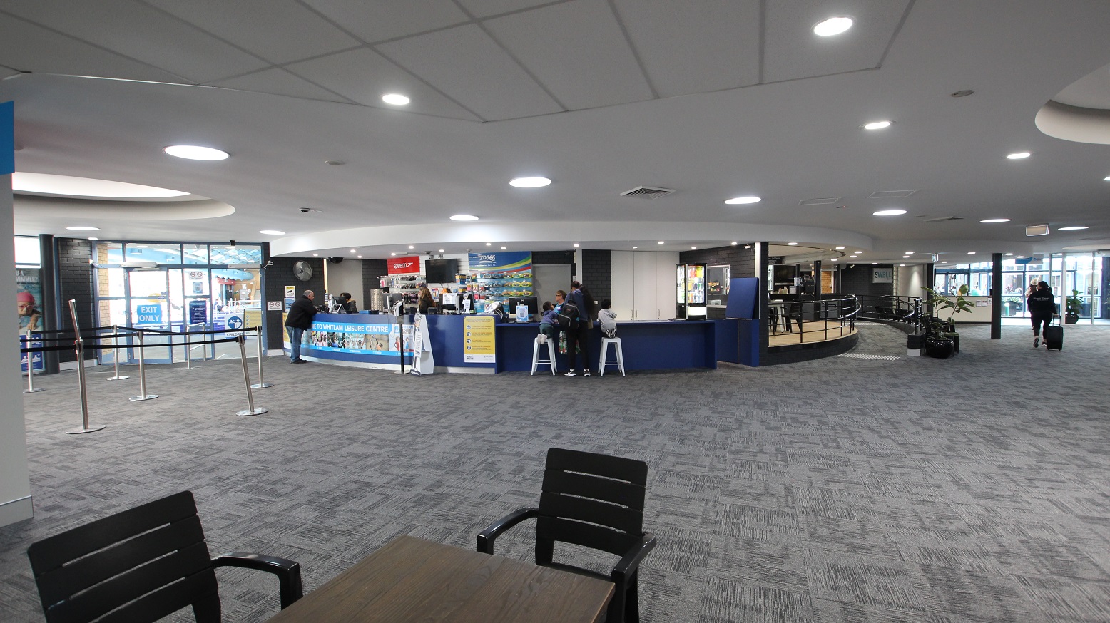View when standing in the seating area containing black chairs and tables opposite of the reception desk. This image includes the peripheral part of the Swell Café with a low ramp-style entry point. The ramp has black metal handrails, and against it is a medium-sized potted plant and black chairs. View when standing in the seating area containing black chairs and tables opposite of the reception desk. This image includes the peripheral part of the Swell Café with a low ramp-style entry point. The ramp has black metal handrails, and against it is a medium-sized potted plant and black chairs.