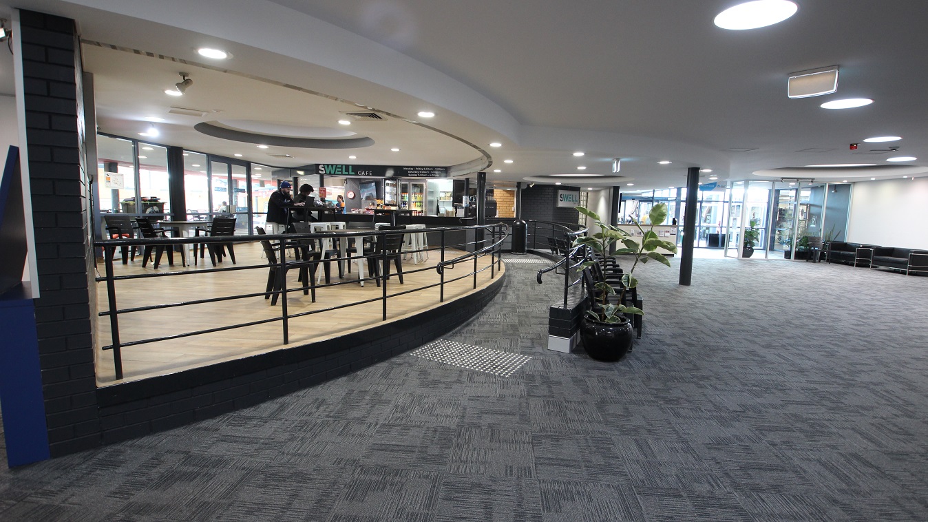 Low-ramp style entrance to the Swell Café. The café is a bit elevated from the ground floor and can be distinguished by its black and white tables & chairs and its walnut wooden floor. On the left side of the café are glass walls and doors which give a natural light coming from the outside. Low-ramp style entrance to the Swell Café. The café is a bit elevated from the ground floor and can be distinguished by its black and white tables & chairs and its walnut wooden floor. On the left side of the café are glass walls and doors which give a natural light coming from the outside.