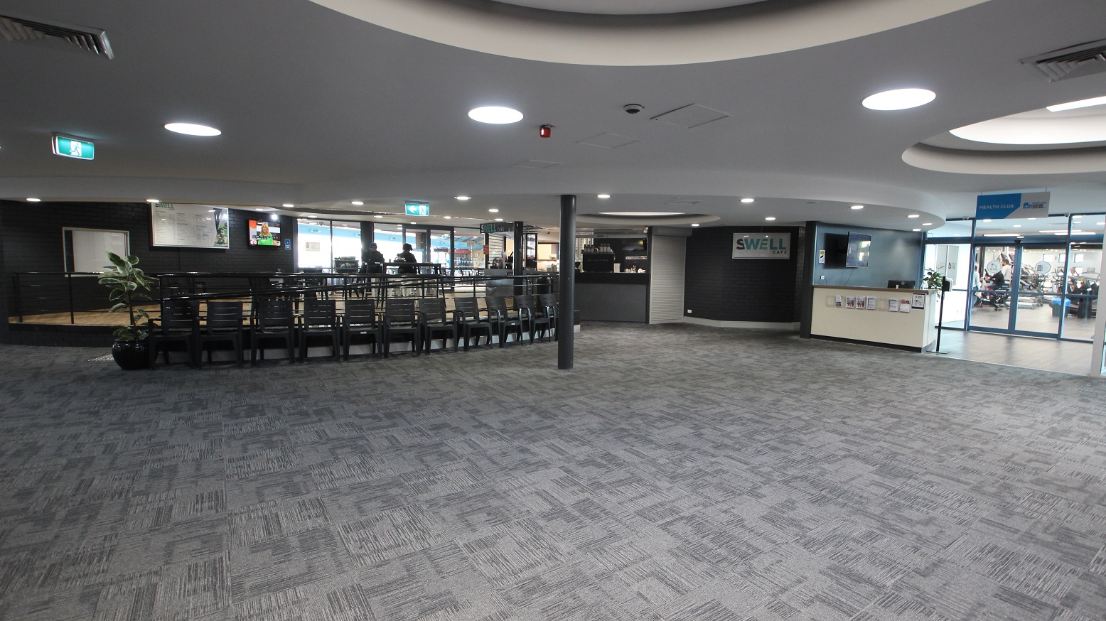 The image shows the Swell Café and Health Club. Just below the low-ramp style entrance of the cafe are several black chairs against the handrails. Heading straight after turning right from the reception desk is the Health Club, which can be identified by its white reception desk and blue wall. It has glass doors that allow guests to see other guests enjoying the Health Club facility. The image shows the Swell Café and Health Club. Just below the low-ramp style entrance of the cafe are several black chairs against the handrails. Heading straight after turning right from the reception desk is the Health Club, which can be identified by its white reception desk and blue wall. It has glass doors that allow guests to see other guests enjoying the Health Club facility.
