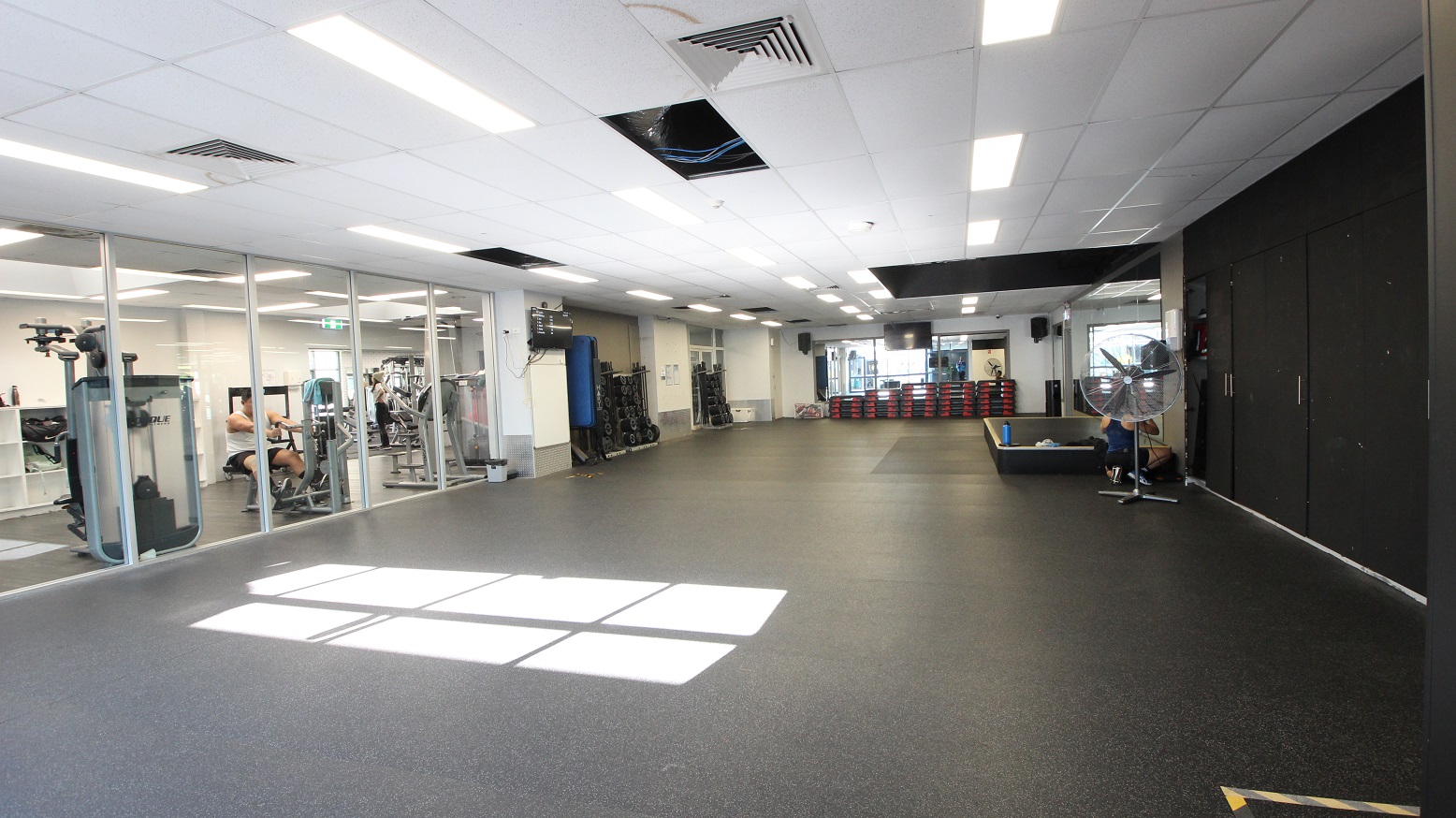 The weight training area, a group exercise studio, and a spin studio at Whitlam Leisure Centre. A large, black rectangular block is placed in the center right after the stand fan. Weight-lifting equipment is arranged against the walls The weight training area, a group exercise studio, and a spin studio at Whitlam Leisure Centre. A large, black rectangular block is placed in the center right after the stand fan. Weight-lifting equipment is arranged against the walls