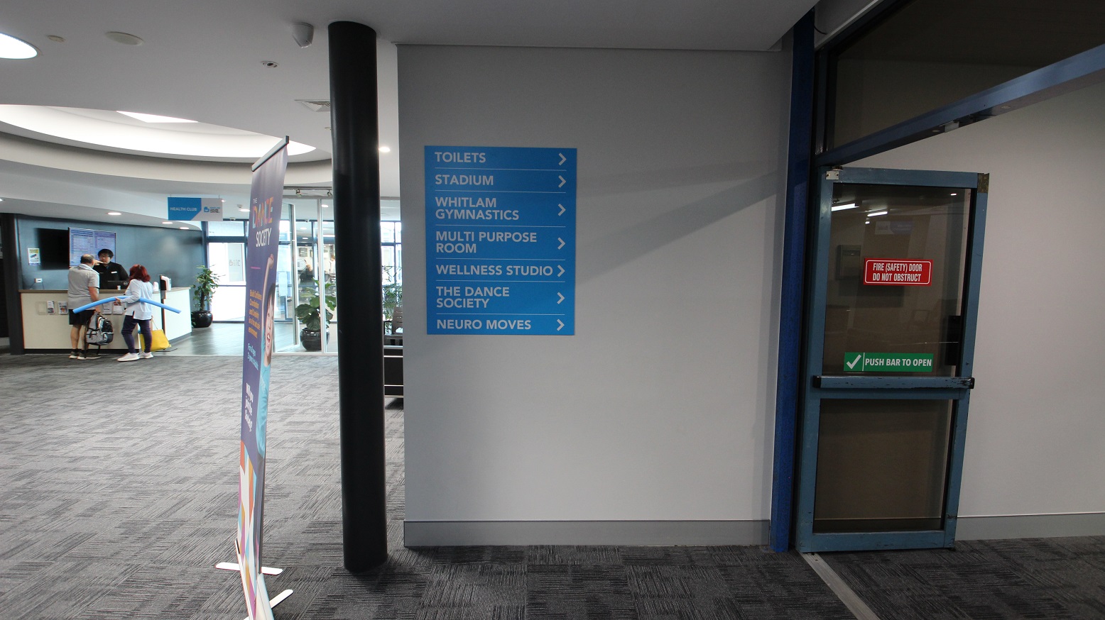 Open, sliding, fire double doors. On the right of these doors is a wall with a class schedule for Bruins Liverpool Skills and Development basketball training and black chairs. There are also a number of chairs settled against the walls at the end of the corridor. Open, sliding, fire double doors. On the right of these doors is a wall with a class schedule for Bruins Liverpool Skills and Development basketball training and black chairs. There are also a number of chairs settled against the walls at the end of the corridor.