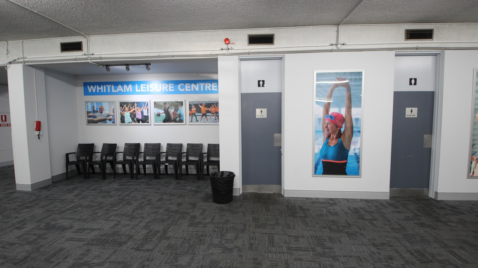 In between the separate toilet rooms for males and females is a photograph of a woman wearing swimming goggles and a swimming cap. Outside the male toilet room is a small rubbish bin. On the left side of the toilet section are black chairs for guests to sit on while waiting for their turn. In between the separate toilet rooms for males and females is a photograph of a woman wearing swimming goggles and a swimming cap. Outside the male toilet room is a small rubbish bin. On the left side of the toilet section are black chairs for guests to sit on while waiting for their turn.