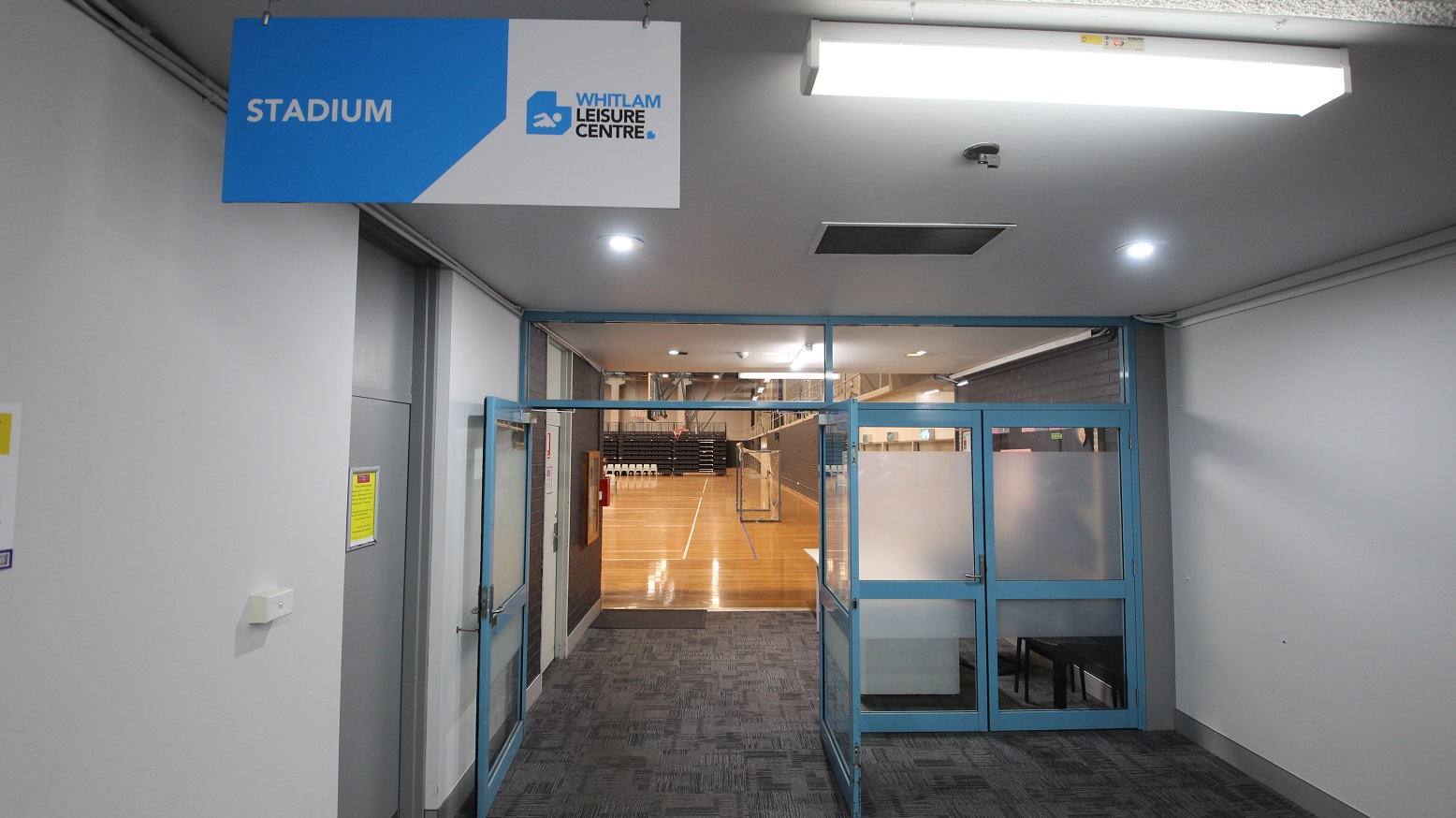 Entrance to the Stadium at Whitlam Leisure Centre. The double door on the left side is open, while the double door with a blurry portion on the right is not. Seen through the open door is the polished timber flooring, with a brown ceiling with dark beams, including fluorescent lig Entrance to the Stadium at Whitlam Leisure Centre. The double door on the left side is open, while the double door with a blurry portion on the right is not. Seen through the open door is the polished timber flooring, with a brown ceiling with dark beams, including fluorescent lig